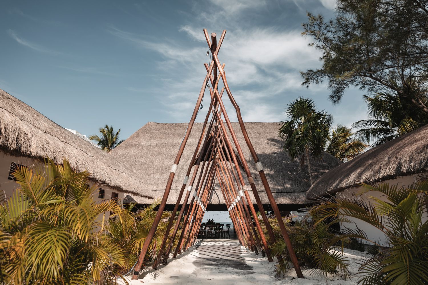 Tropical resort entrance with wooden teepee structure, thatched roof bungalows, and lush palm trees under a blue sky.
