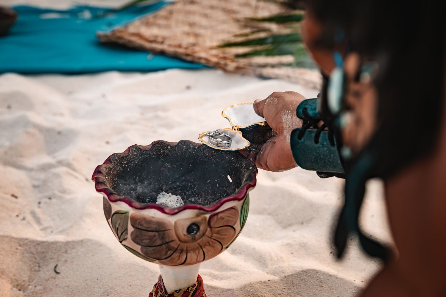 Person pouring liquid into ornate ceremonial bowl on sandy beach during traditional ritual.