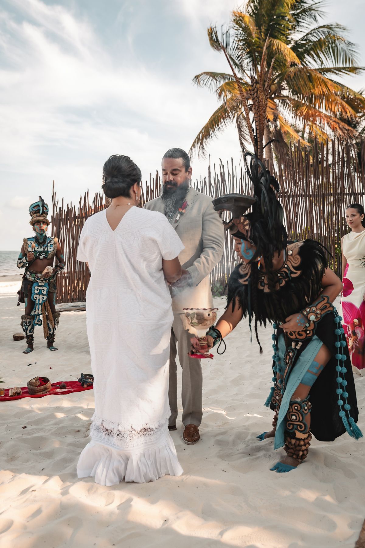 Couple exchanges vows in Mayan beach wedding ceremony with costumed performers in traditional Aztec attire on sandy shore.