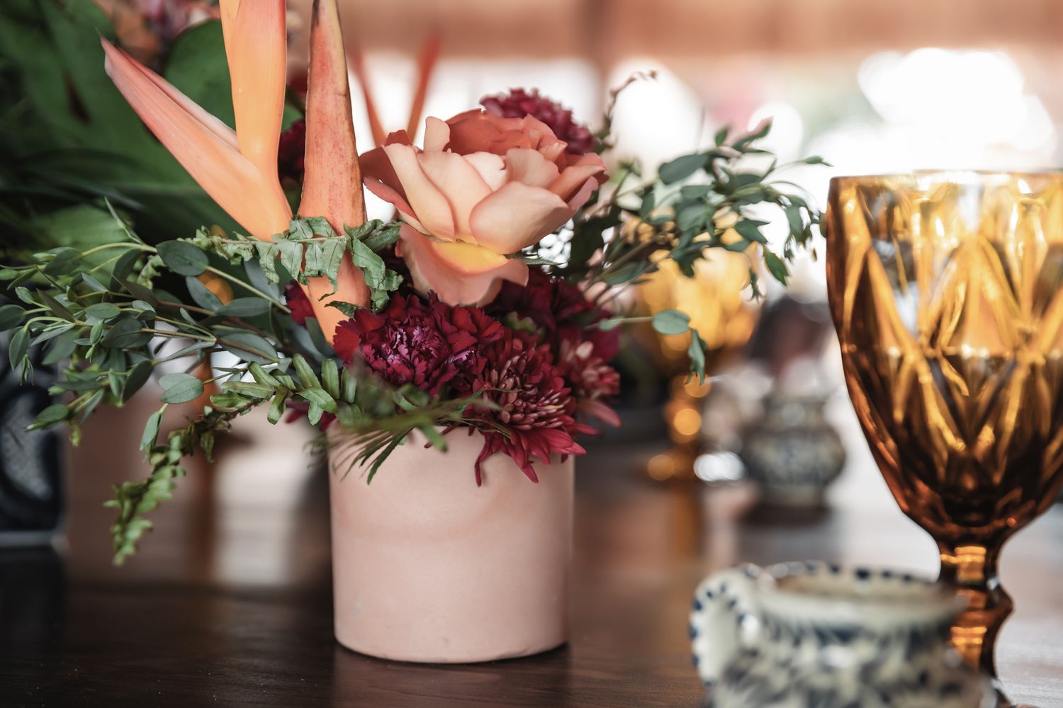 Pink vase with rose, burgundy flowers, and greenery beside an amber cut glass on a wooden table.