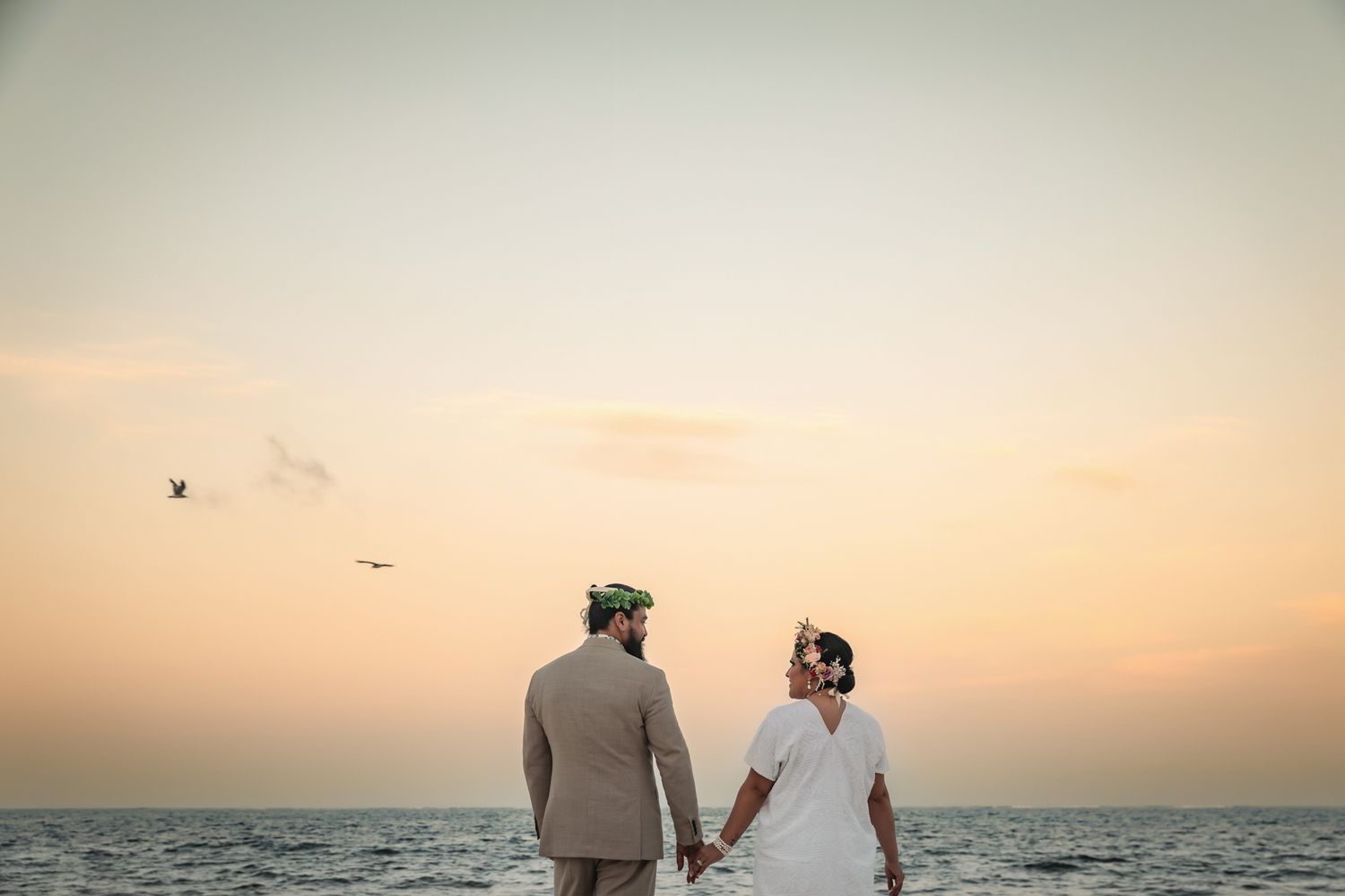 Couple holding hands at sunset beach, bride in white dress and groom in tan suit, ocean horizon backdrop.