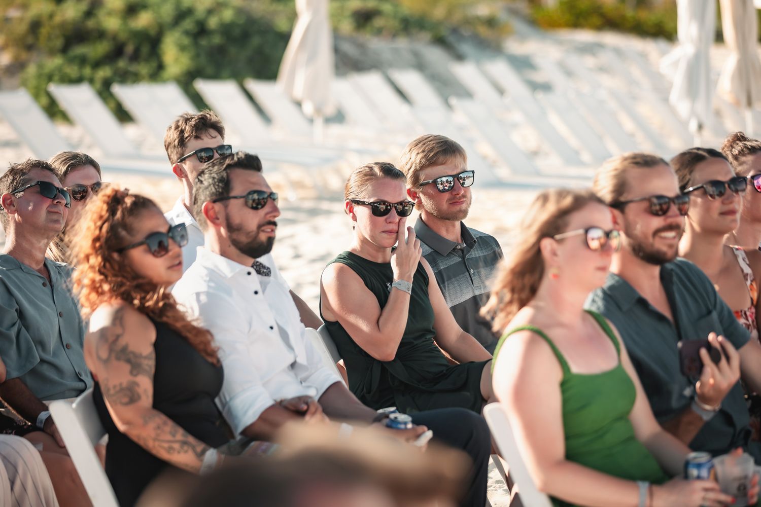 Guests wearing sunglasses seated outdoors at a beach wedding ceremony on a sunny day.