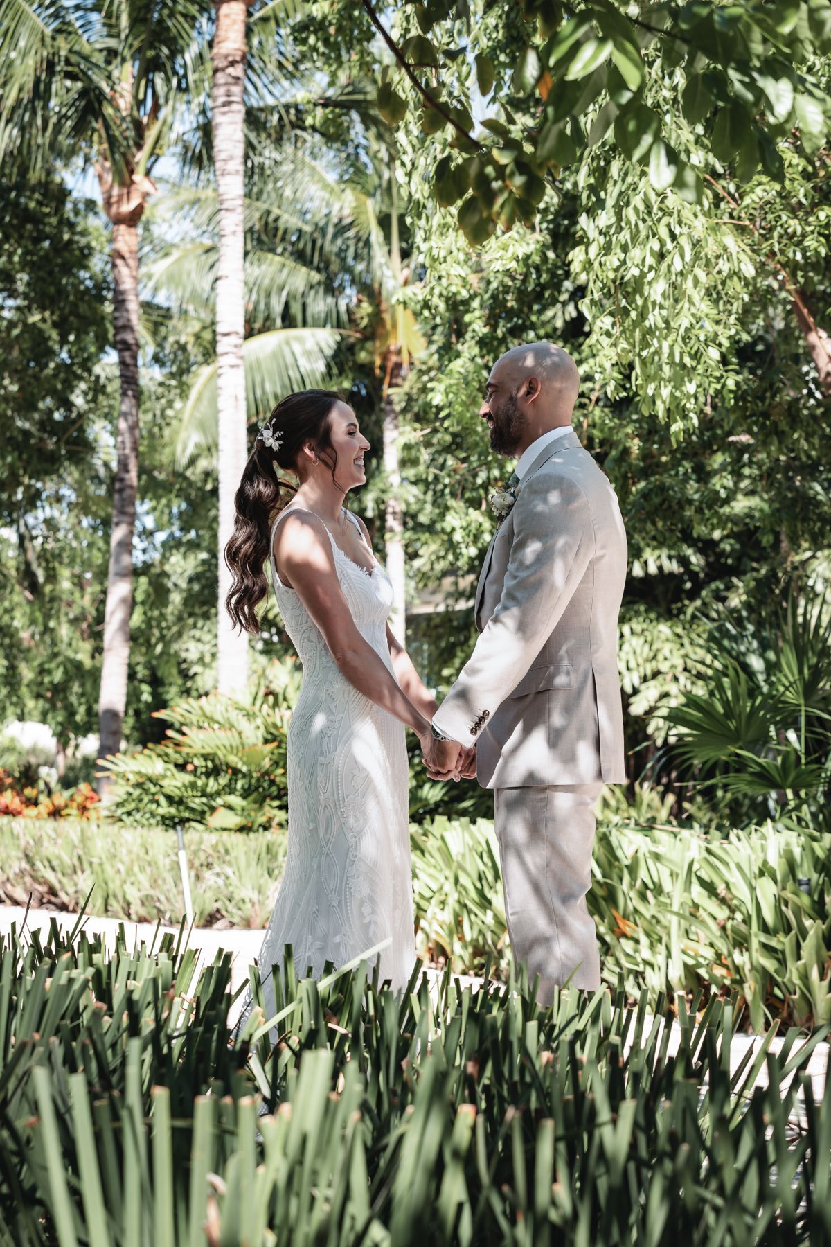 A bride and groom hold hands in a lush tropical garden, she in a white gown, he in a tan suit.