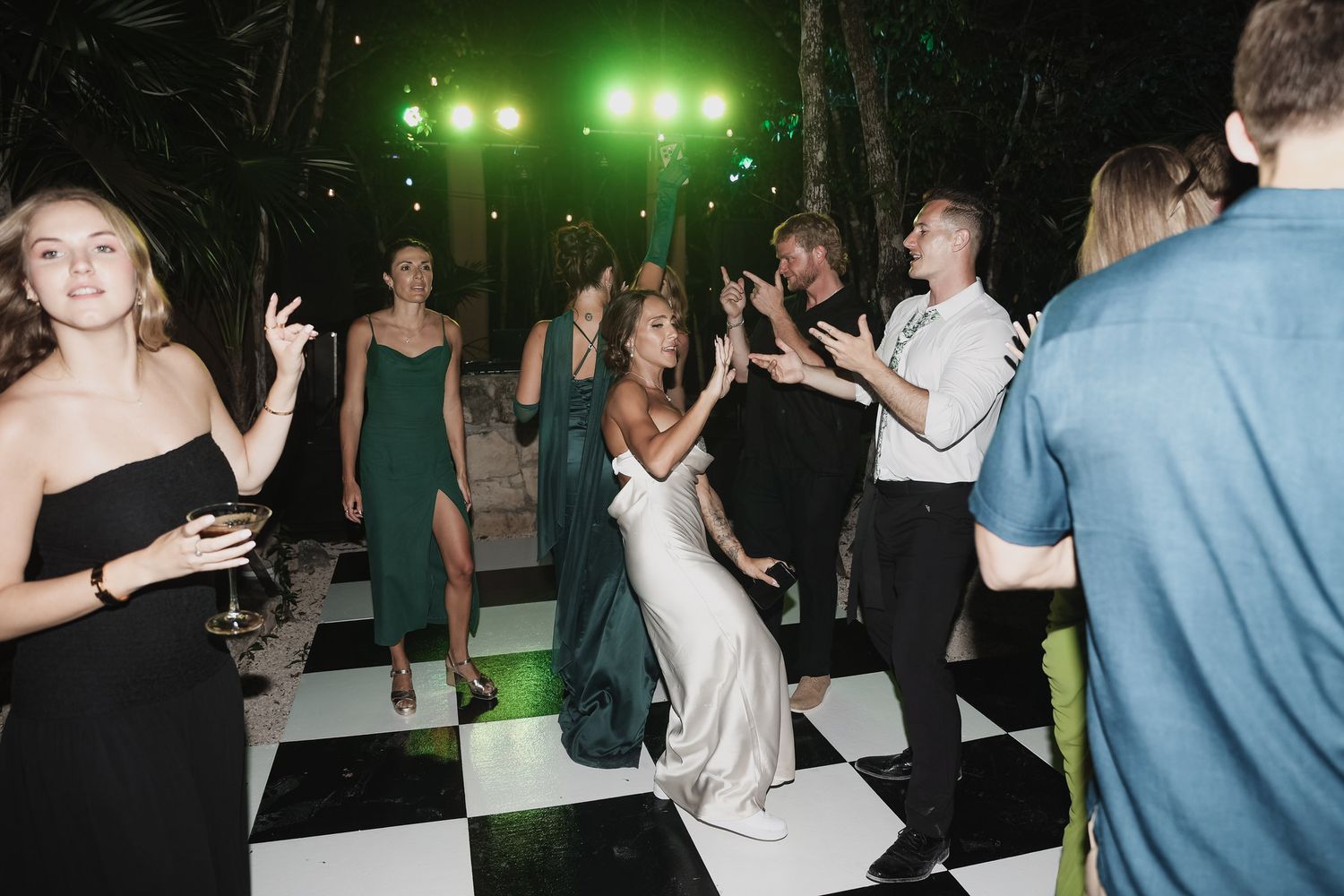 Bride in white gown dancing joyfully on checkered dance floor surrounded by guests at elegant wedding reception.