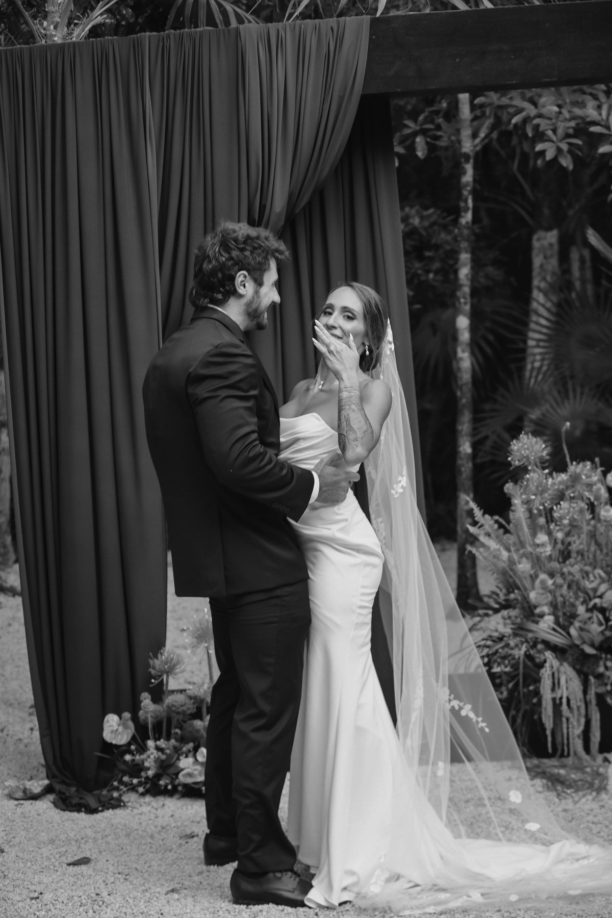 Black and white wedding photo of bride and groom sharing an emotional moment at outdoor altar with draped curtains.