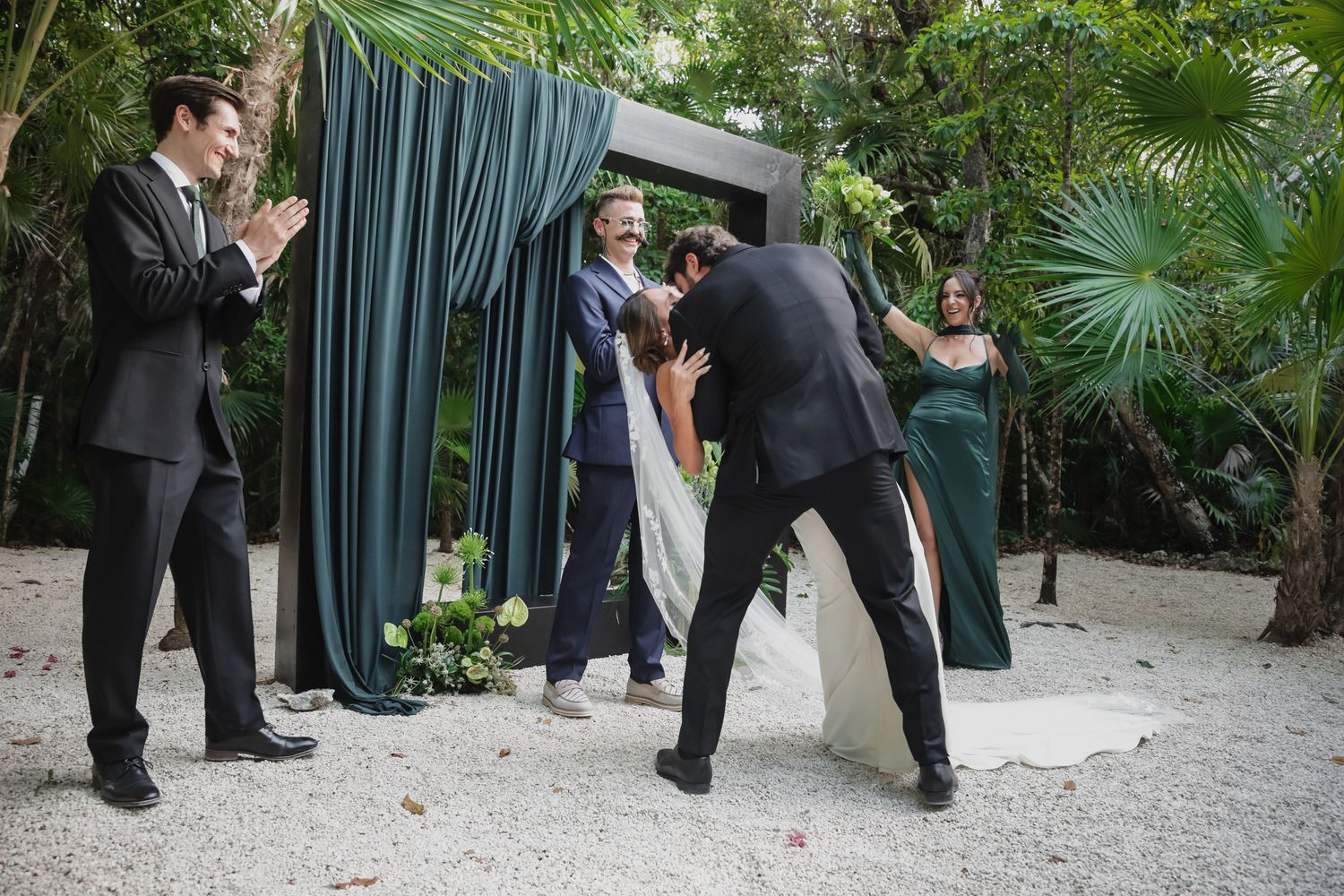 Outdoor wedding ceremony in tropical garden with green draped arch, bride and groom sharing first kiss with wedding party.