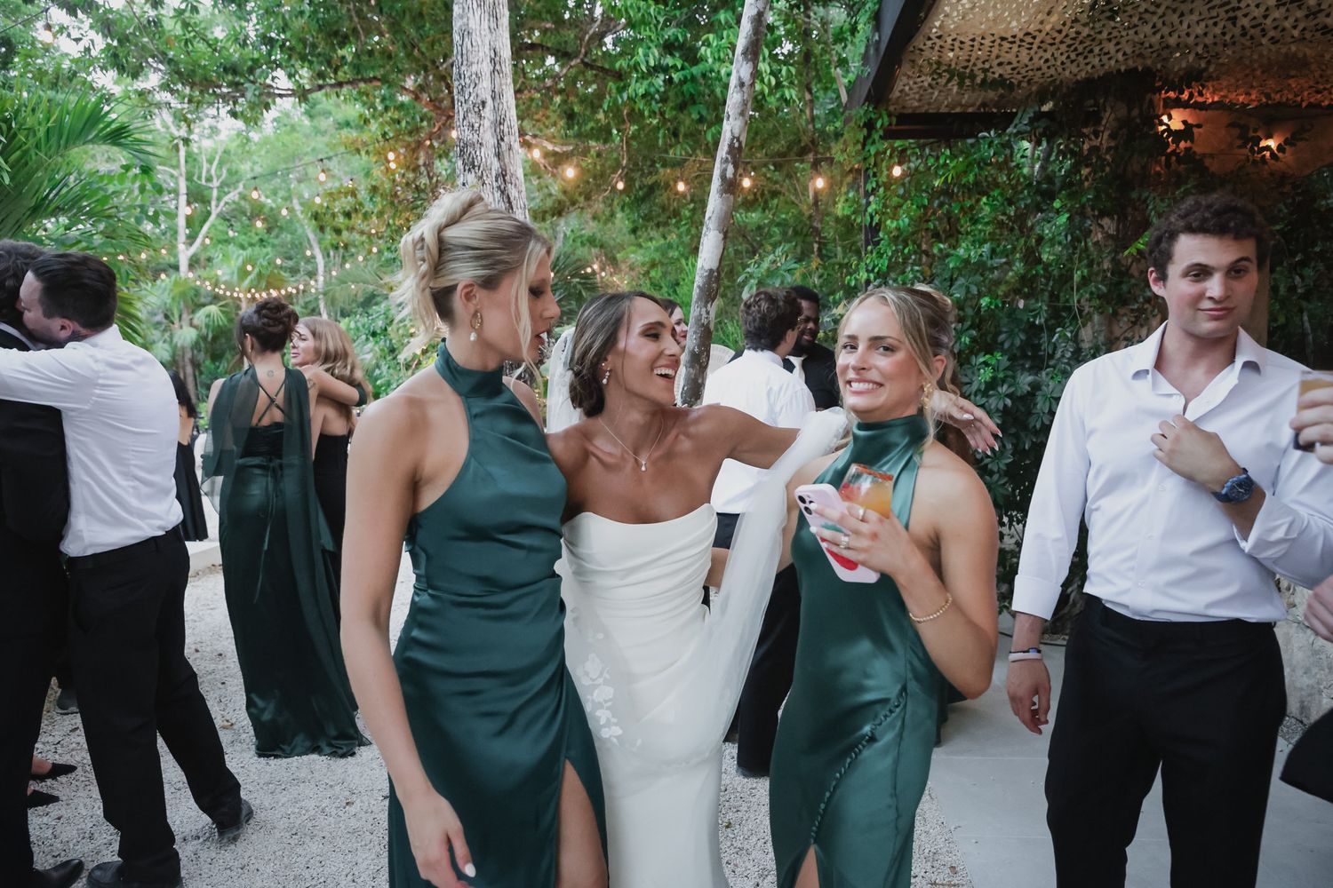 Bride in white gown laughing with bridesmaids in emerald green dresses at outdoor tropical wedding reception.