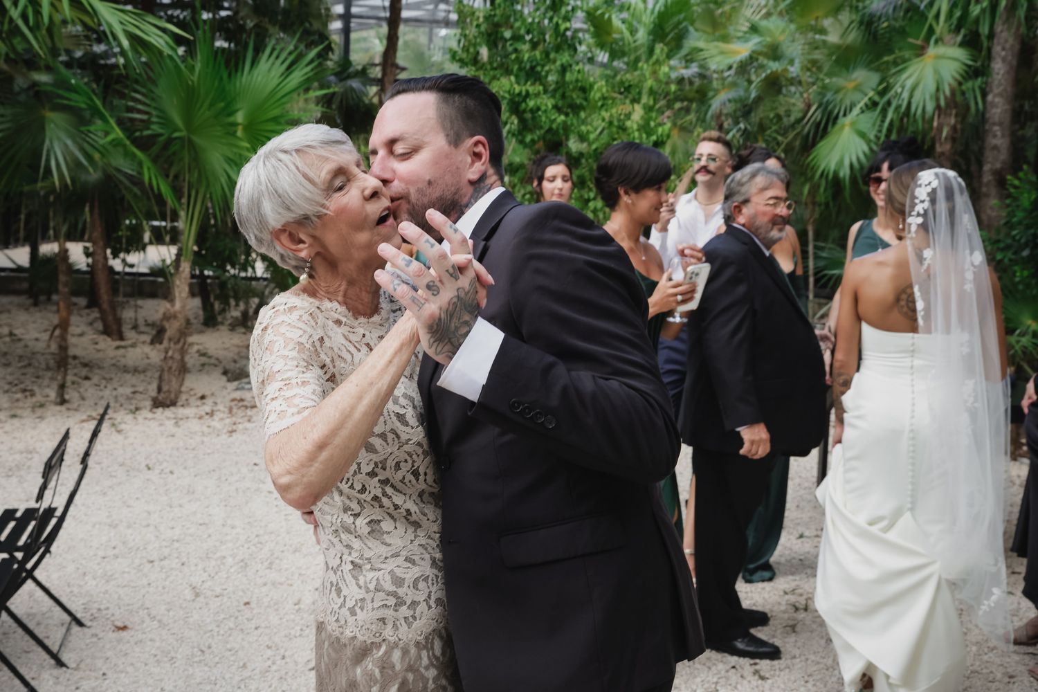 Groom shares a tender kiss with elderly woman at outdoor wedding ceremony with bride visible in background.