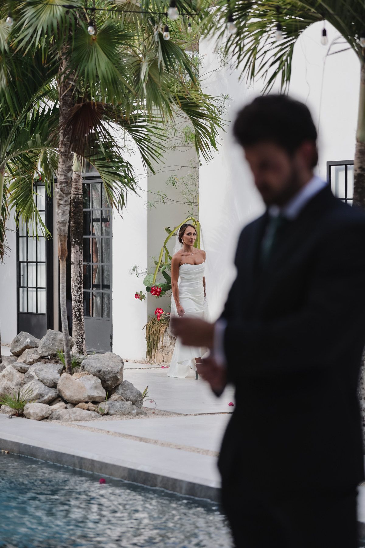 Bride in strapless white gown holding bouquet near pool with palm trees as groom stands in foreground.