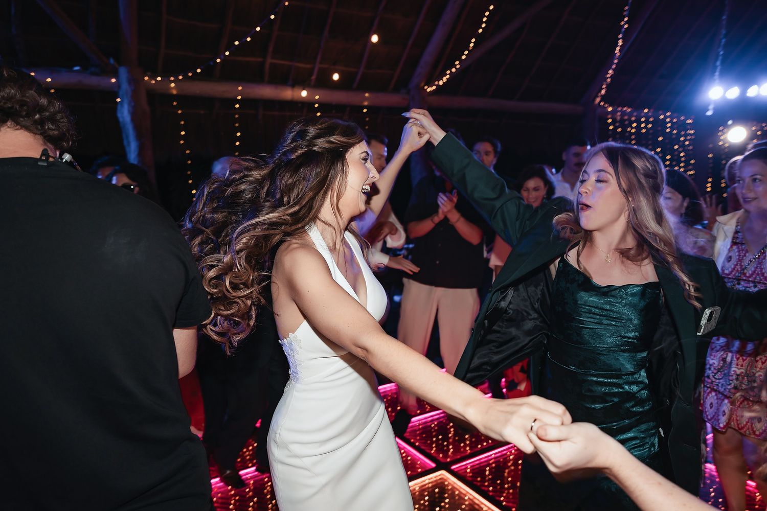 Bride in white dress dancing energetically with guests at a festive wedding reception with string lights.