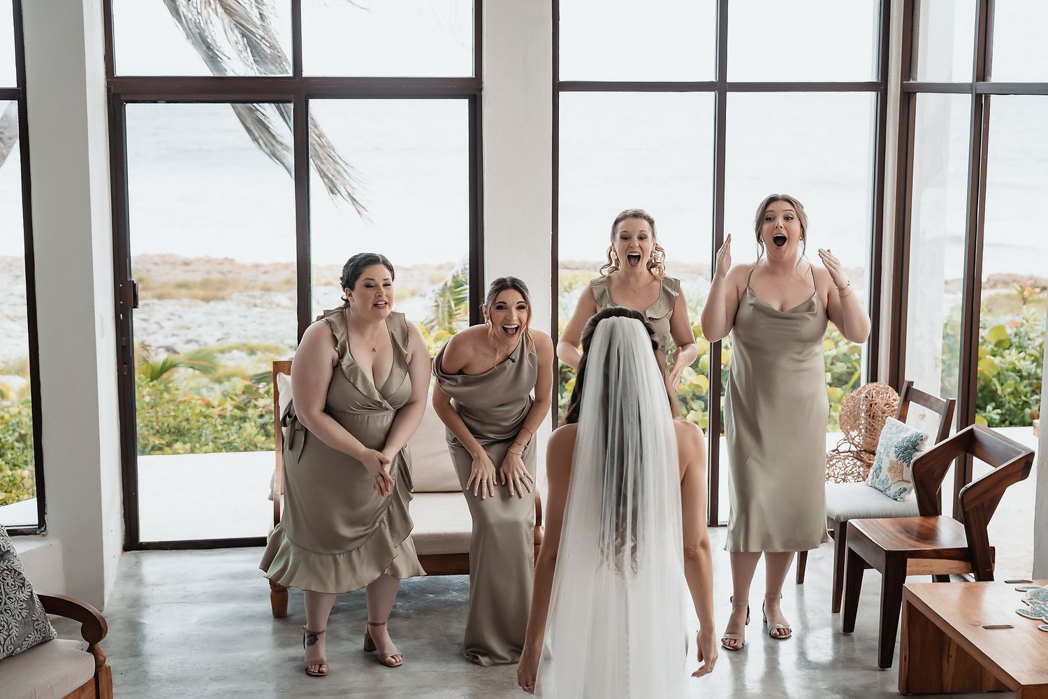 Four bridesmaids in sage dresses react with joy and excitement seeing the bride in her wedding veil.