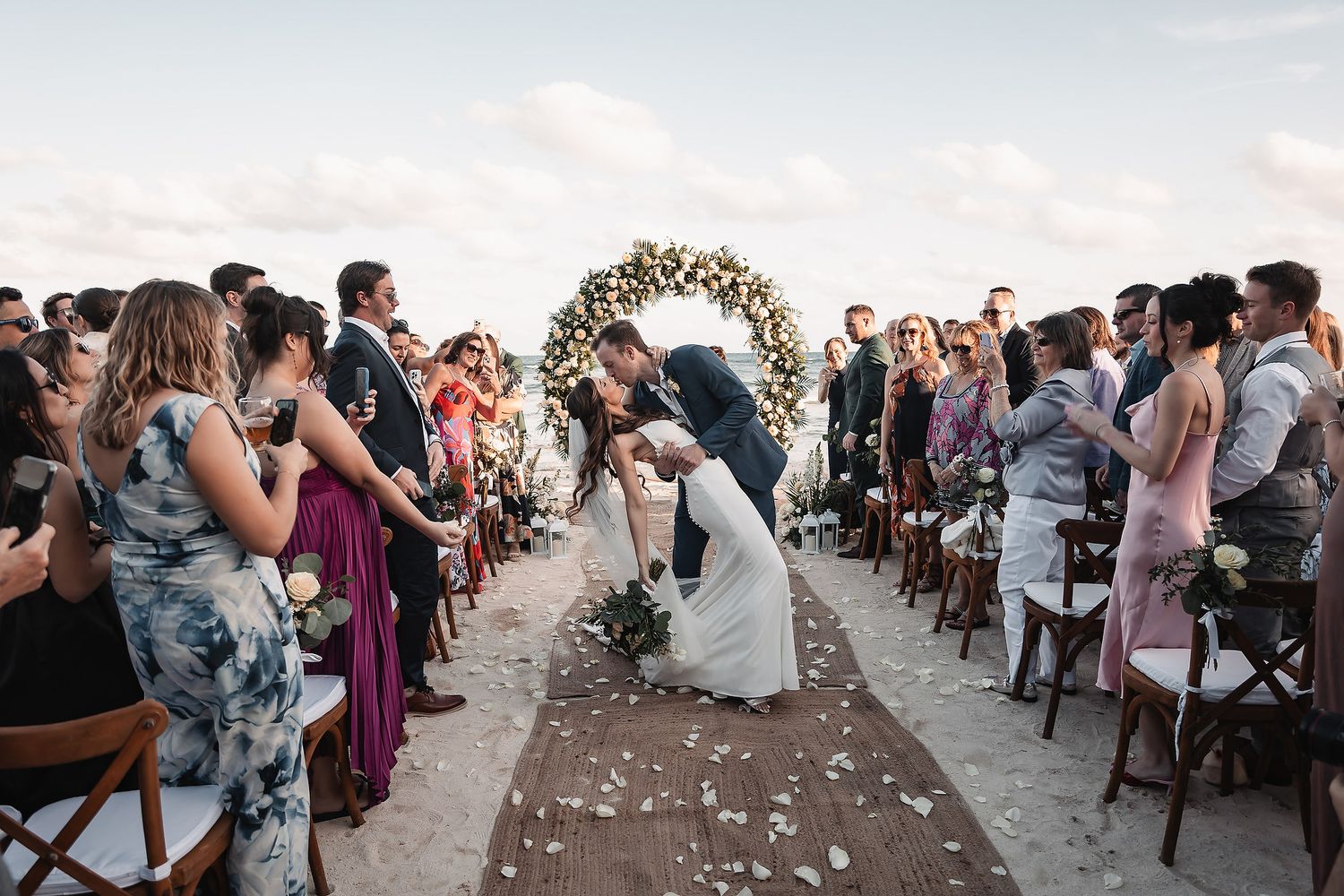 Couple sharing a romantic dip kiss at beach wedding ceremony with floral arch and guests cheering.