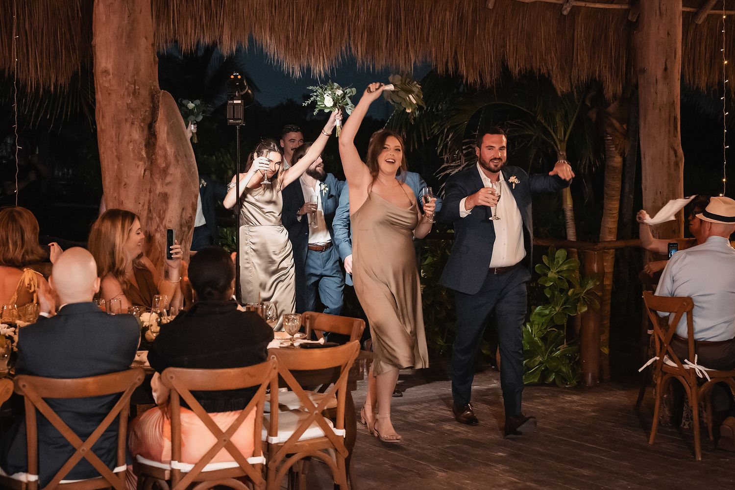 Wedding party makes grand entrance at tropical outdoor reception venue with thatched roof and lush greenery.