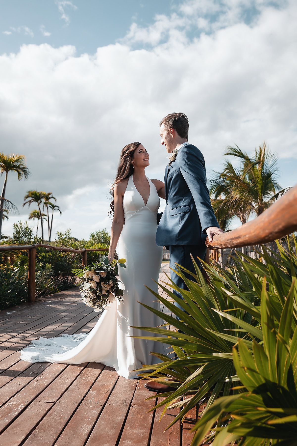 Bride in elegant white gown and groom in blue suit laugh together on a sunny tropical resort deck with palm trees.