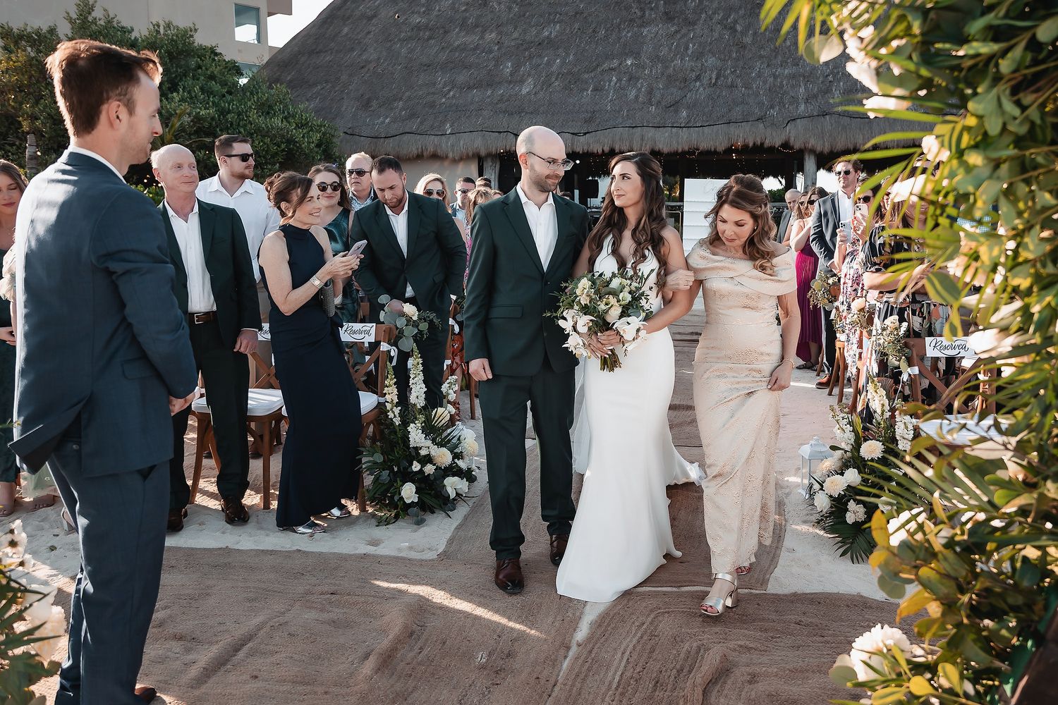 Bride in white gown walks down outdoor aisle with escort at elegant destination wedding ceremony.