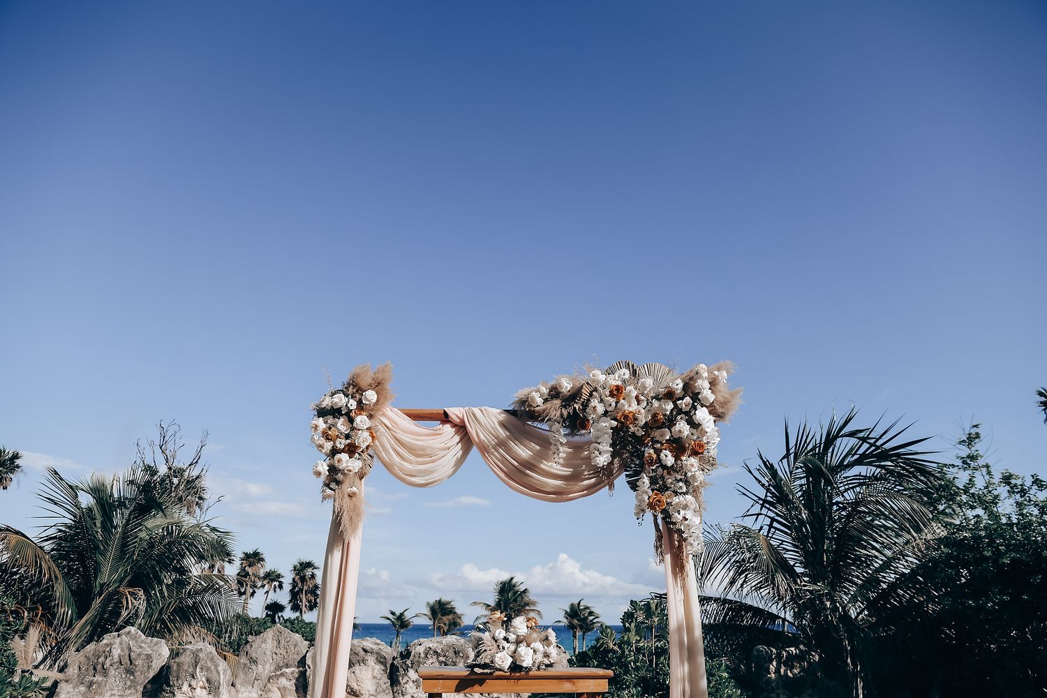 A beachfront wedding ceremony setup with white fabric draped archway and floral decorations against blue ocean backdrop.
