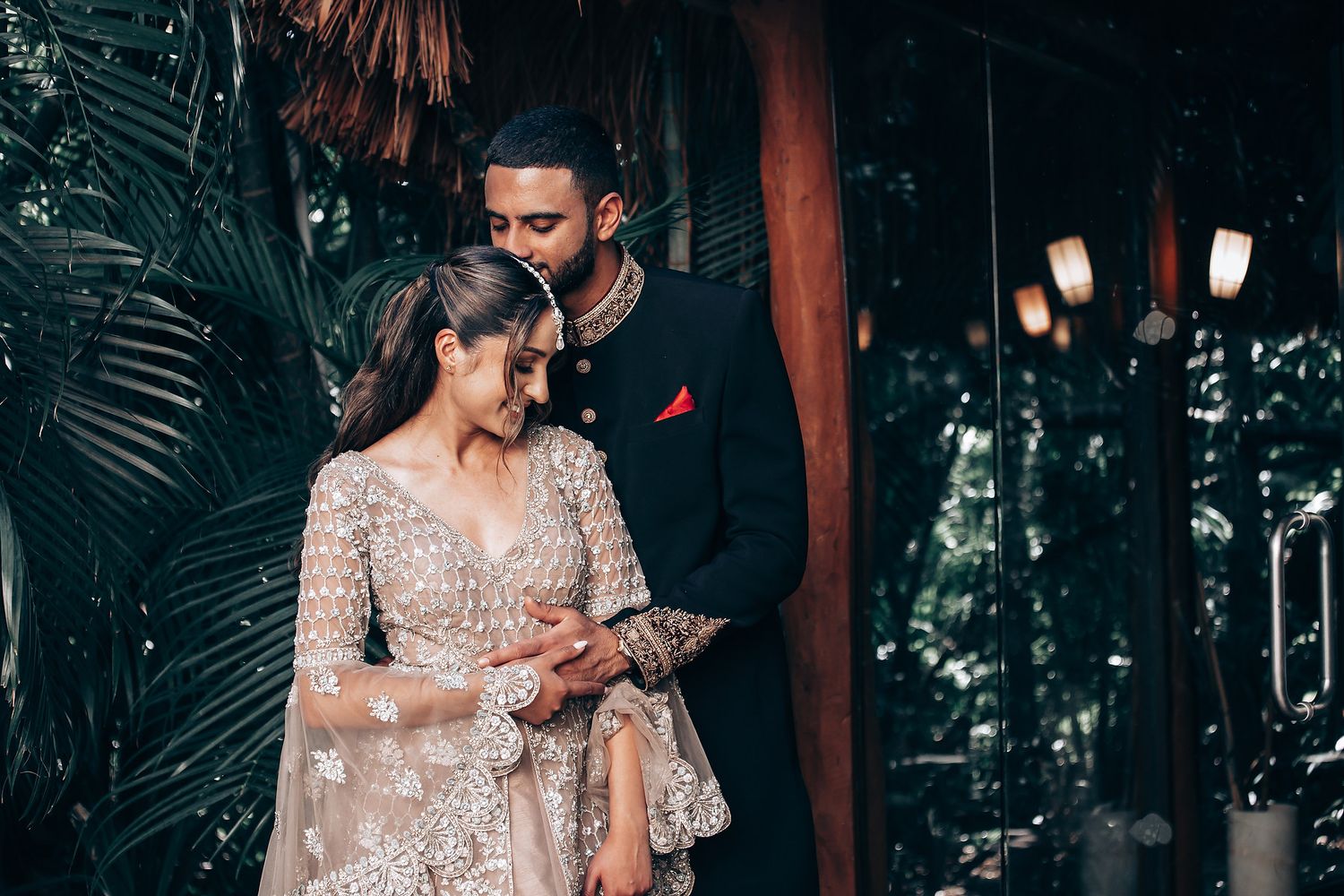 Couple in ornate wedding attire standing on wooden walkway surrounded by tropical foliage.