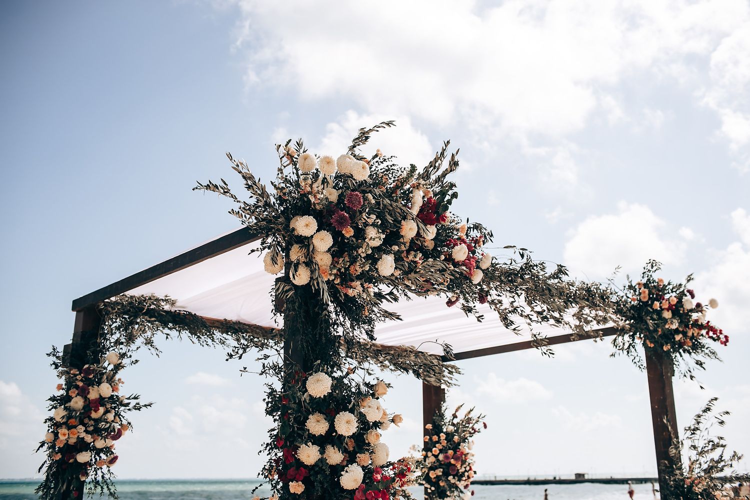 Beachfront wedding ceremony setup with floral arch and white chairs against blue sky.