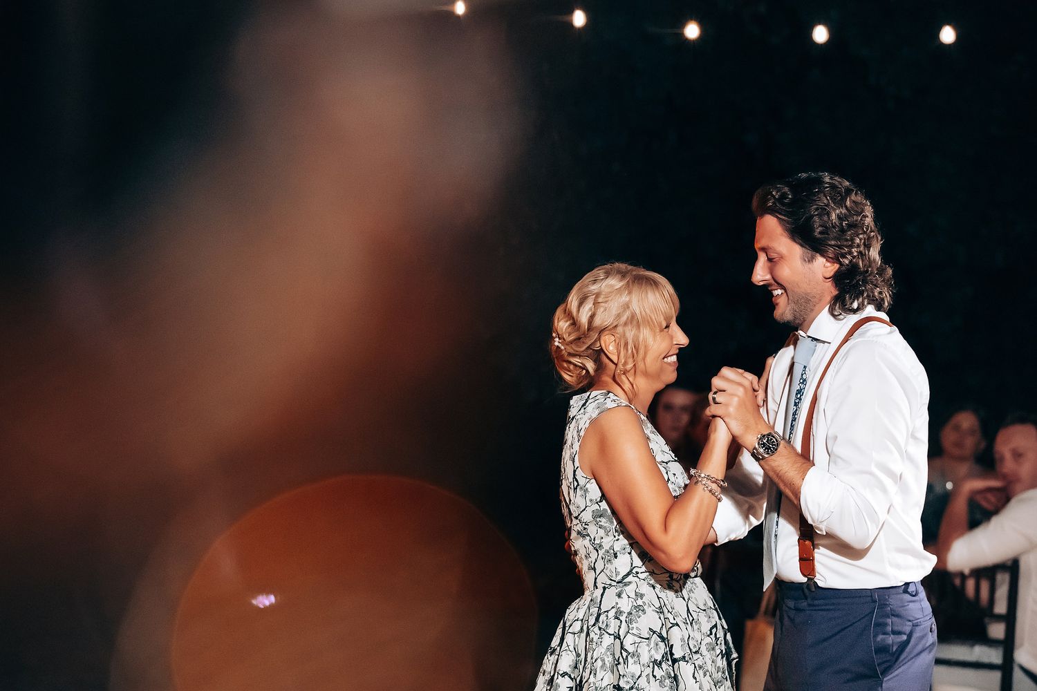 Wedding guests dance together under string lights at an outdoor evening reception celebration.