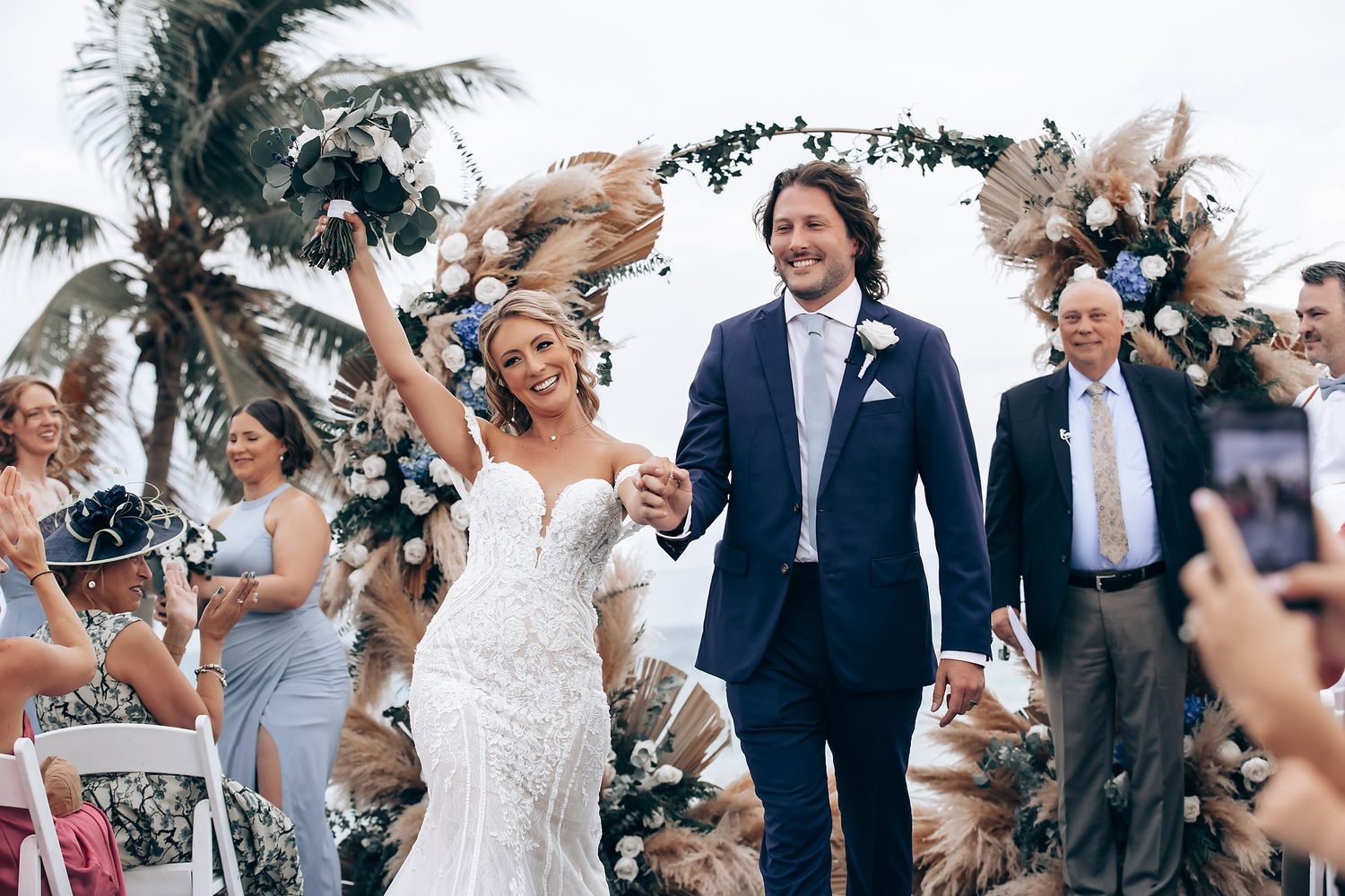 Newlyweds celebrate while walking down the aisle under a tropical arch decorated with pampas grass.