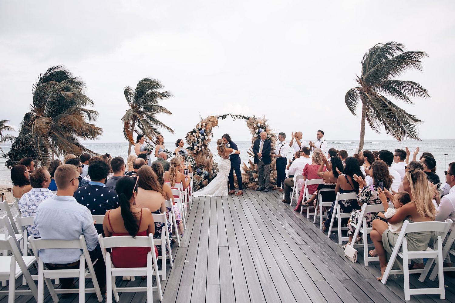 Beachfront wedding ceremony setup with white chairs and swaying palm trees against an overcast sky.