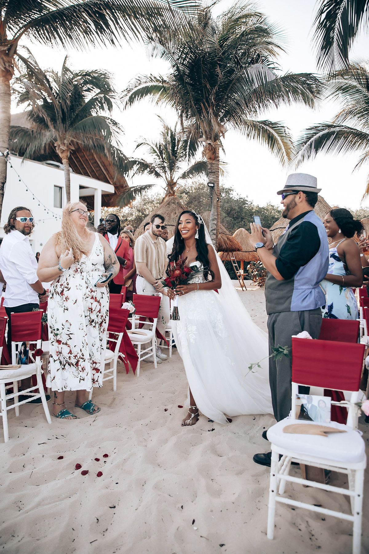 Beach wedding ceremony setup with white chairs and burgundy accents against palm tree backdrop.