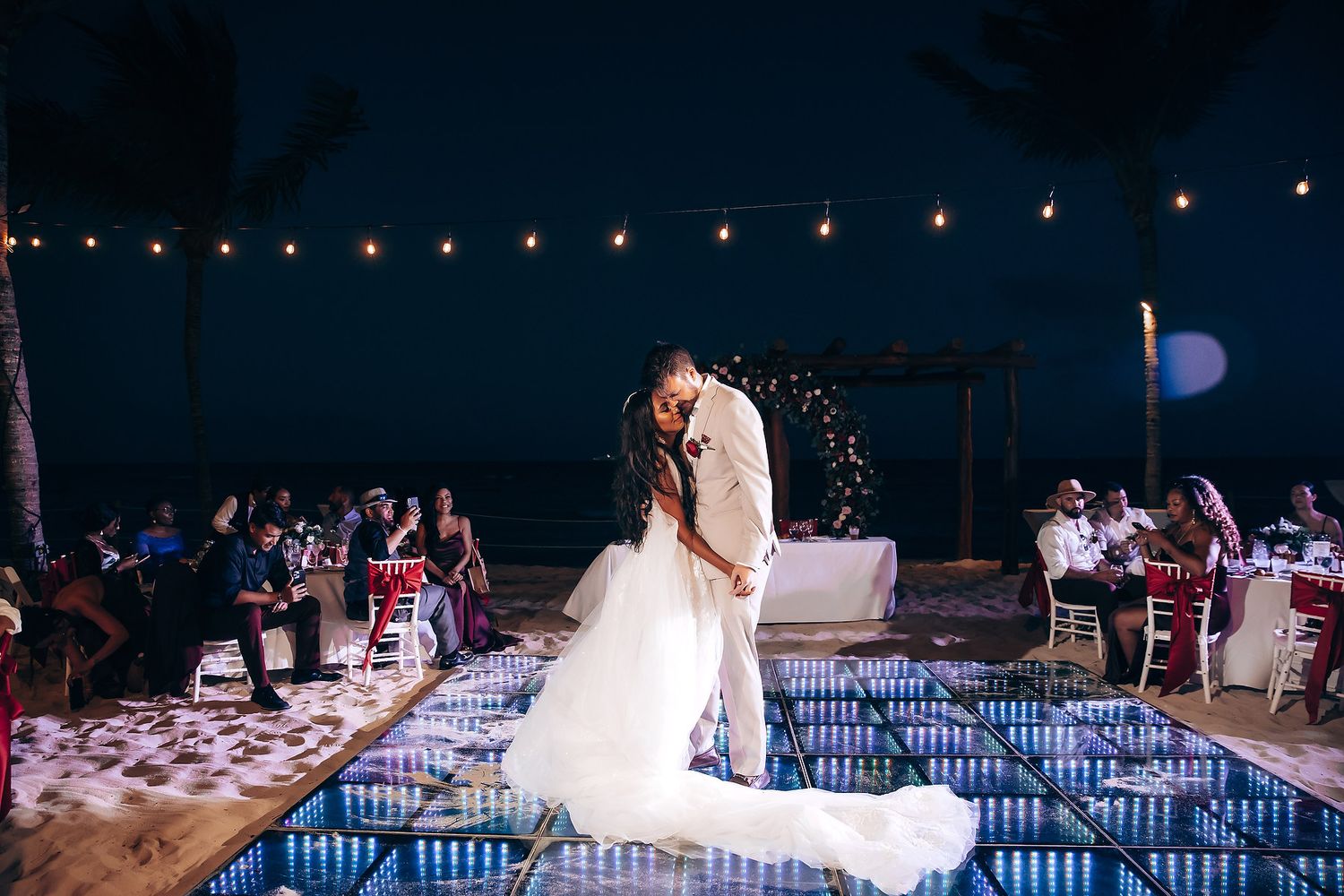 Evening beach wedding reception with string lights and illuminated dance floor setup.