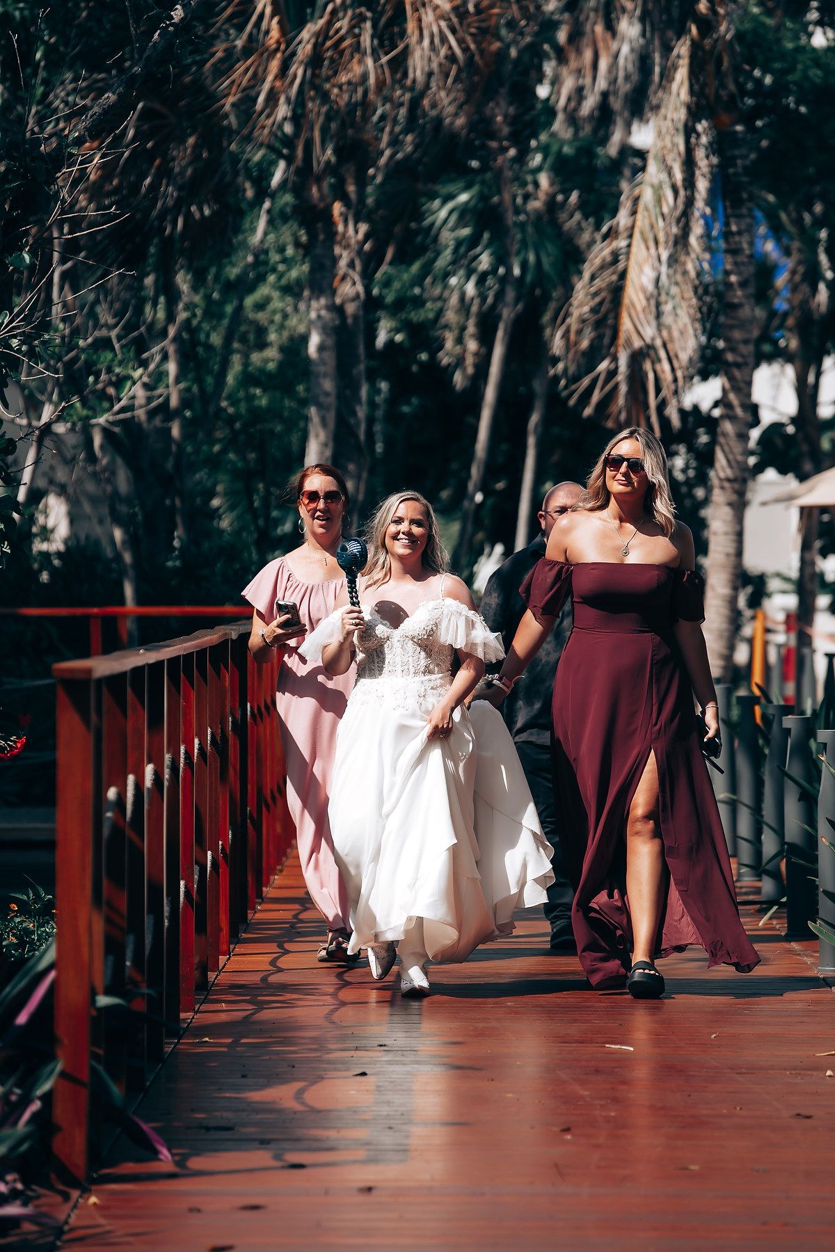 A wedding party walks across a red wooden bridge surrounded by trees on a sunny day.