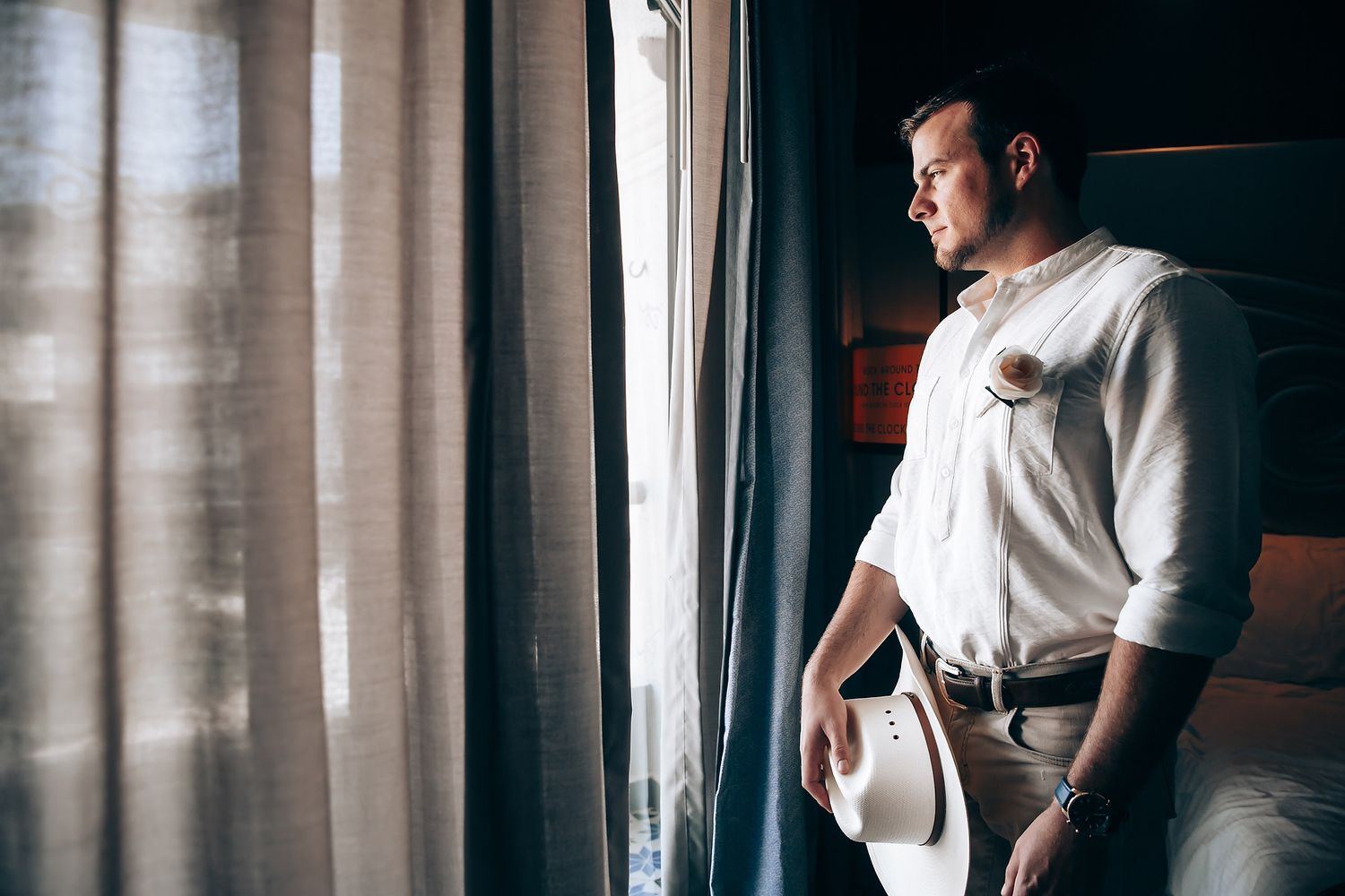 A person in white stands thoughtfully by a window with sheer curtains.
