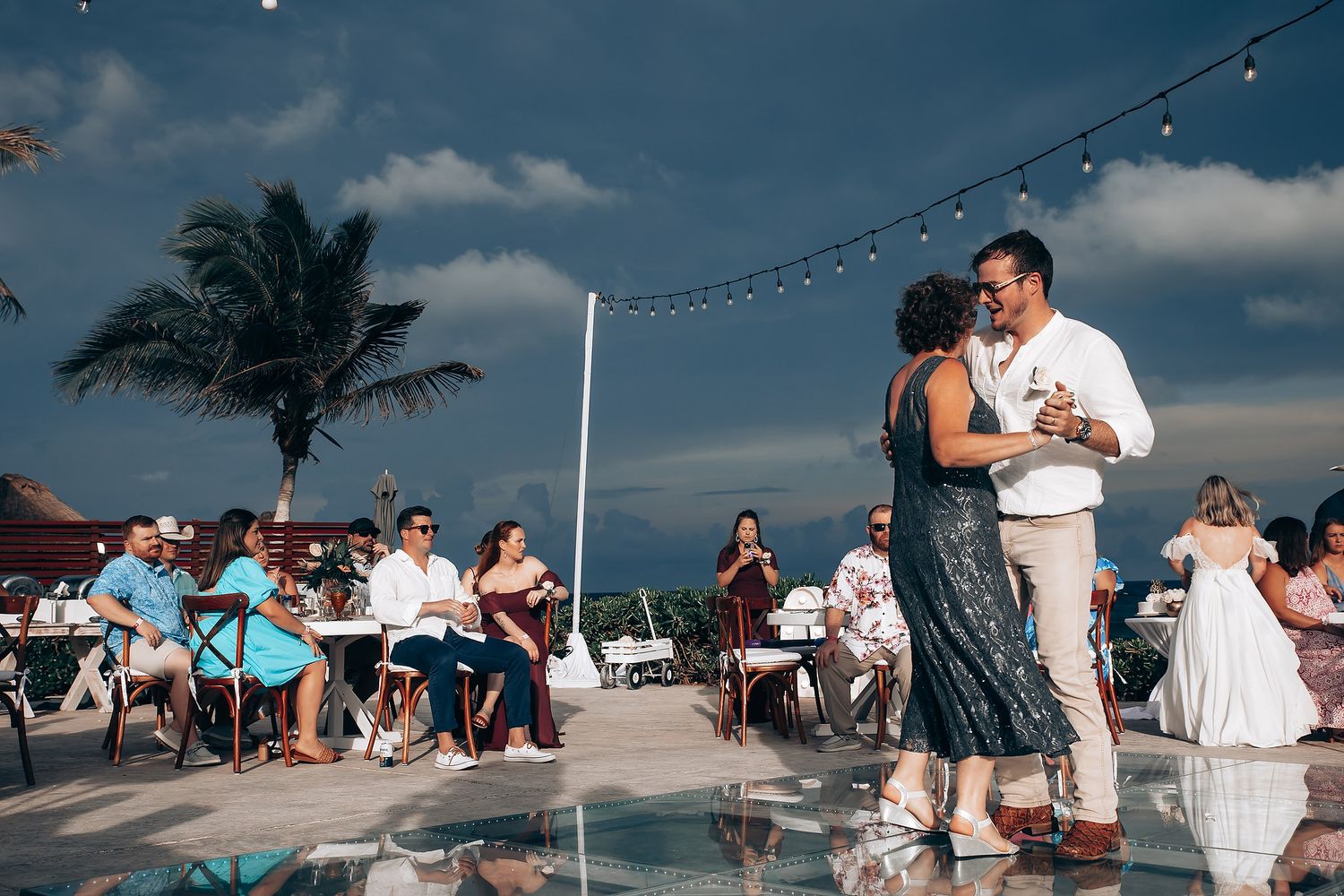 Couple dancing on an outdoor patio with string lights and palm trees during a tropical wedding reception.