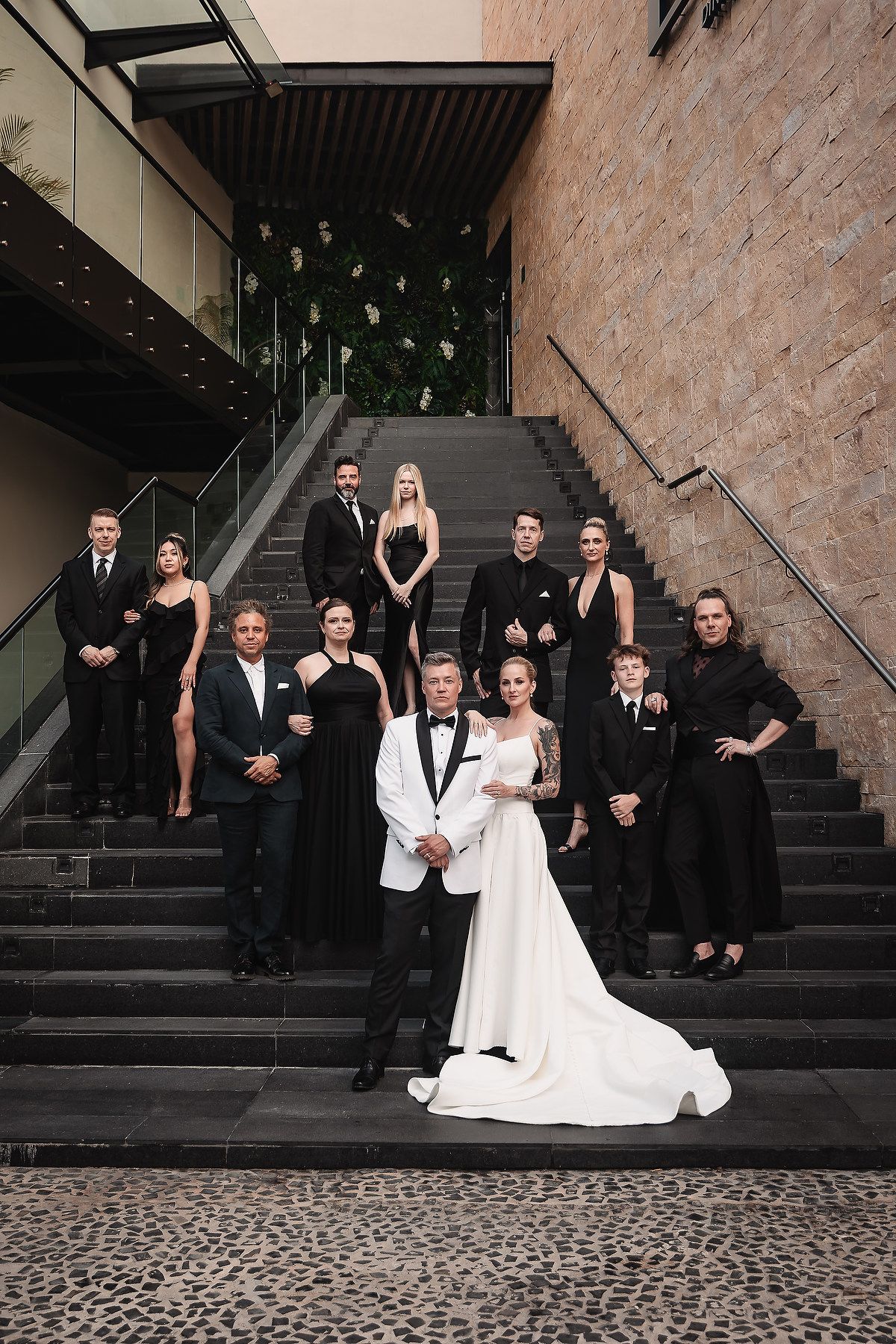 Wedding party in black attire poses dramatically on stone steps with brick wall backdrop.