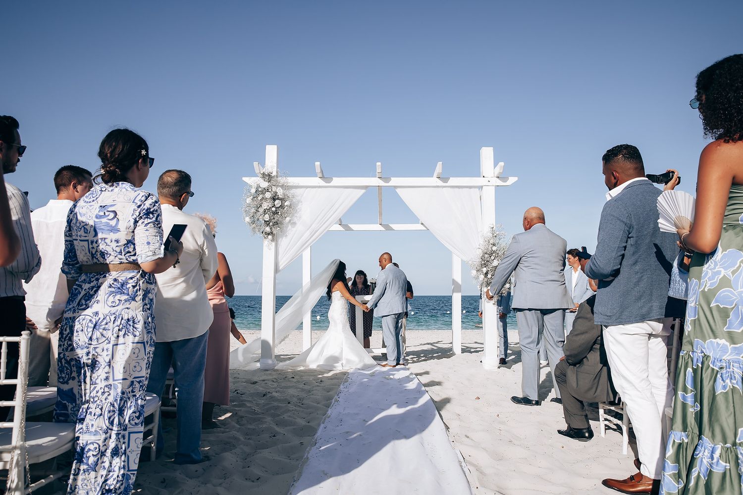 Beachside wedding setup with white pergola and chairs against bright blue sky.