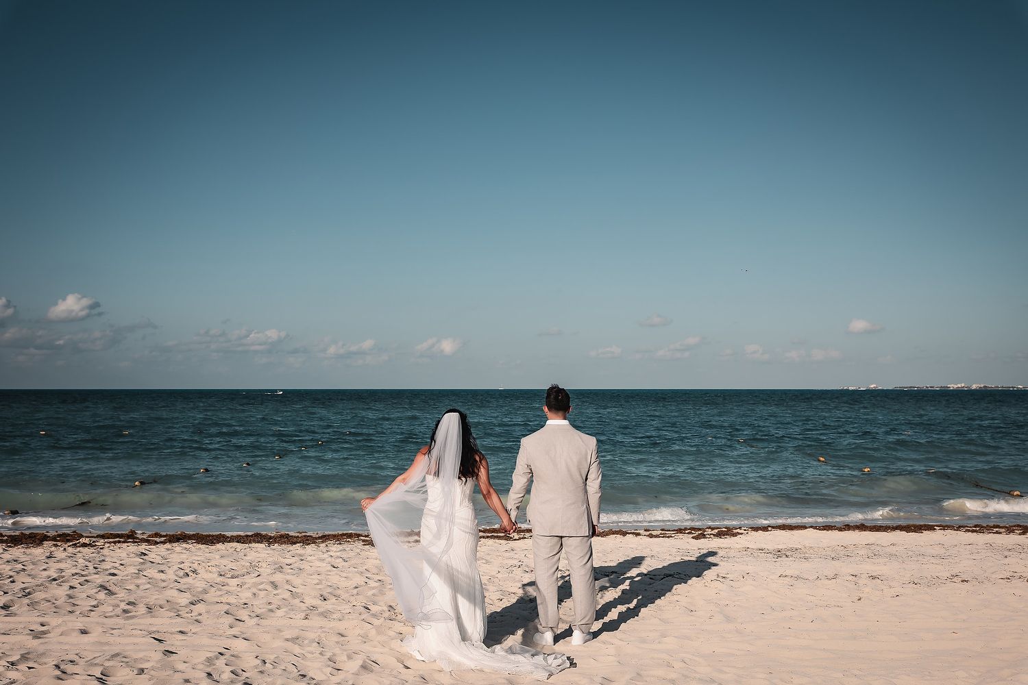 A couple in wedding attire walks hand in hand on a tropical beach with turquoise ocean and blue sky in the background.