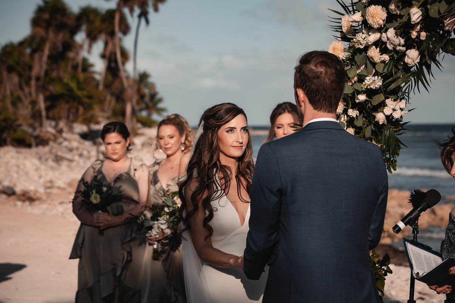 Wedding ceremony in progress on sandy beach with floral arch and ocean backdrop.