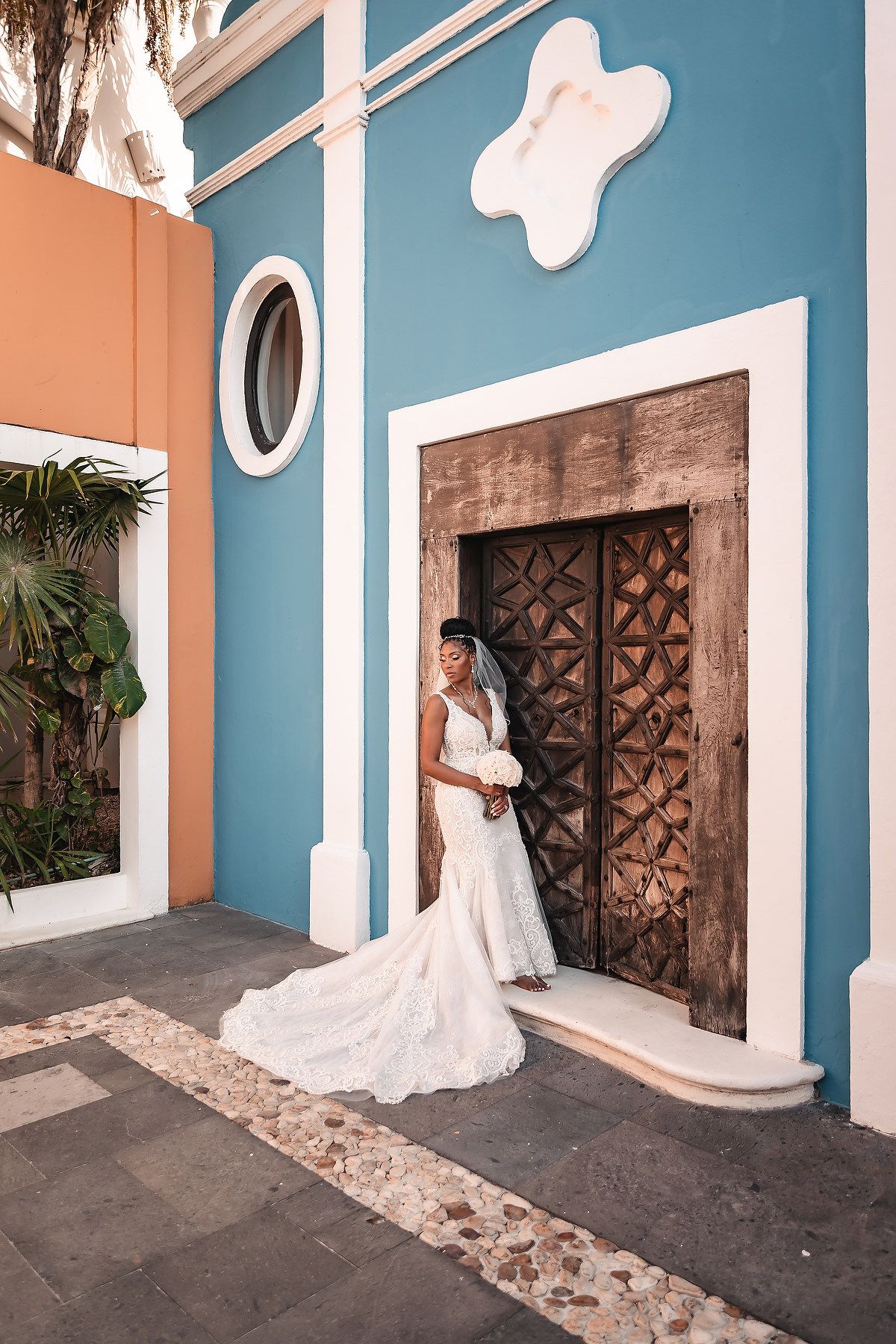 A person in a wedding gown poses against a blue and coral wall with an ornate wooden door.