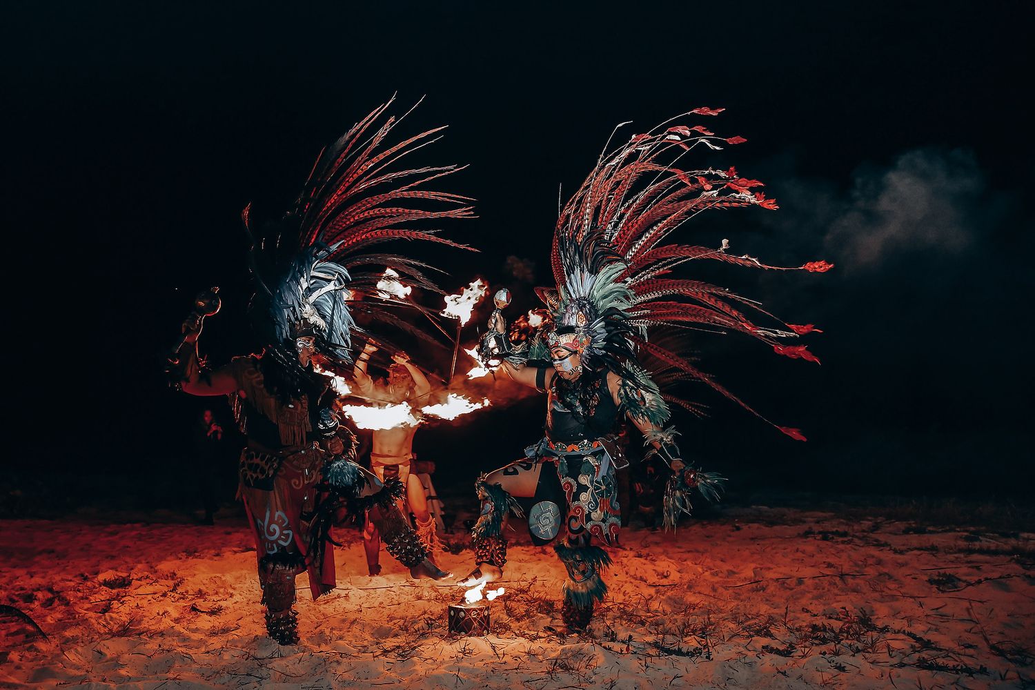 Traditional dancers in elaborate feathered headdresses perform an ancient ritual dance against a dark red backdrop.