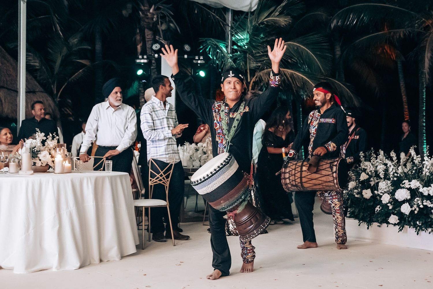 Wedding guests wave their hands in celebration during an indoor reception with floral decorations.