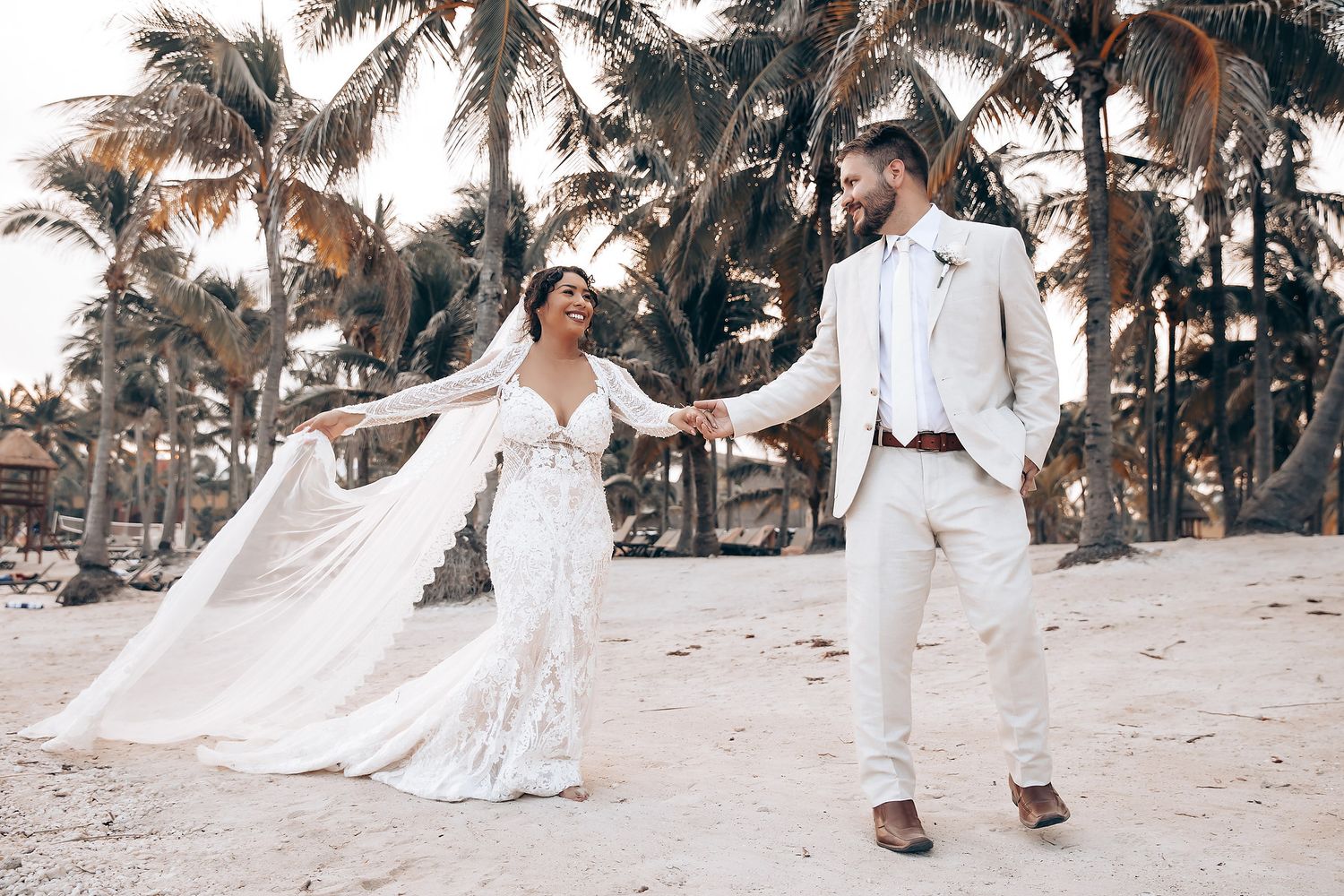 A couple in wedding attire poses together on a tropical beach with palm trees in the background.