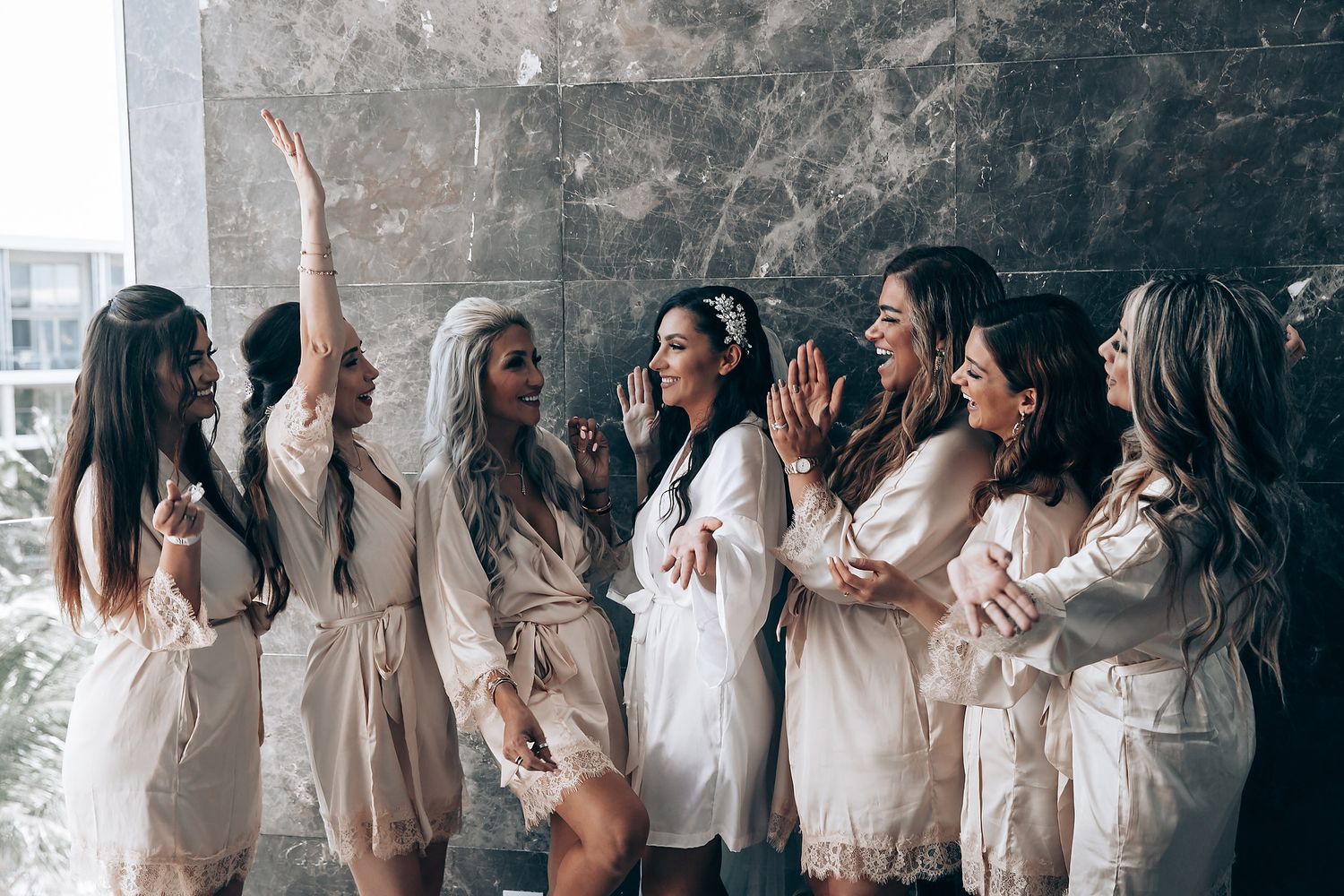 A group of women in matching beige silk robes celebrate together by a window during a special occasion.