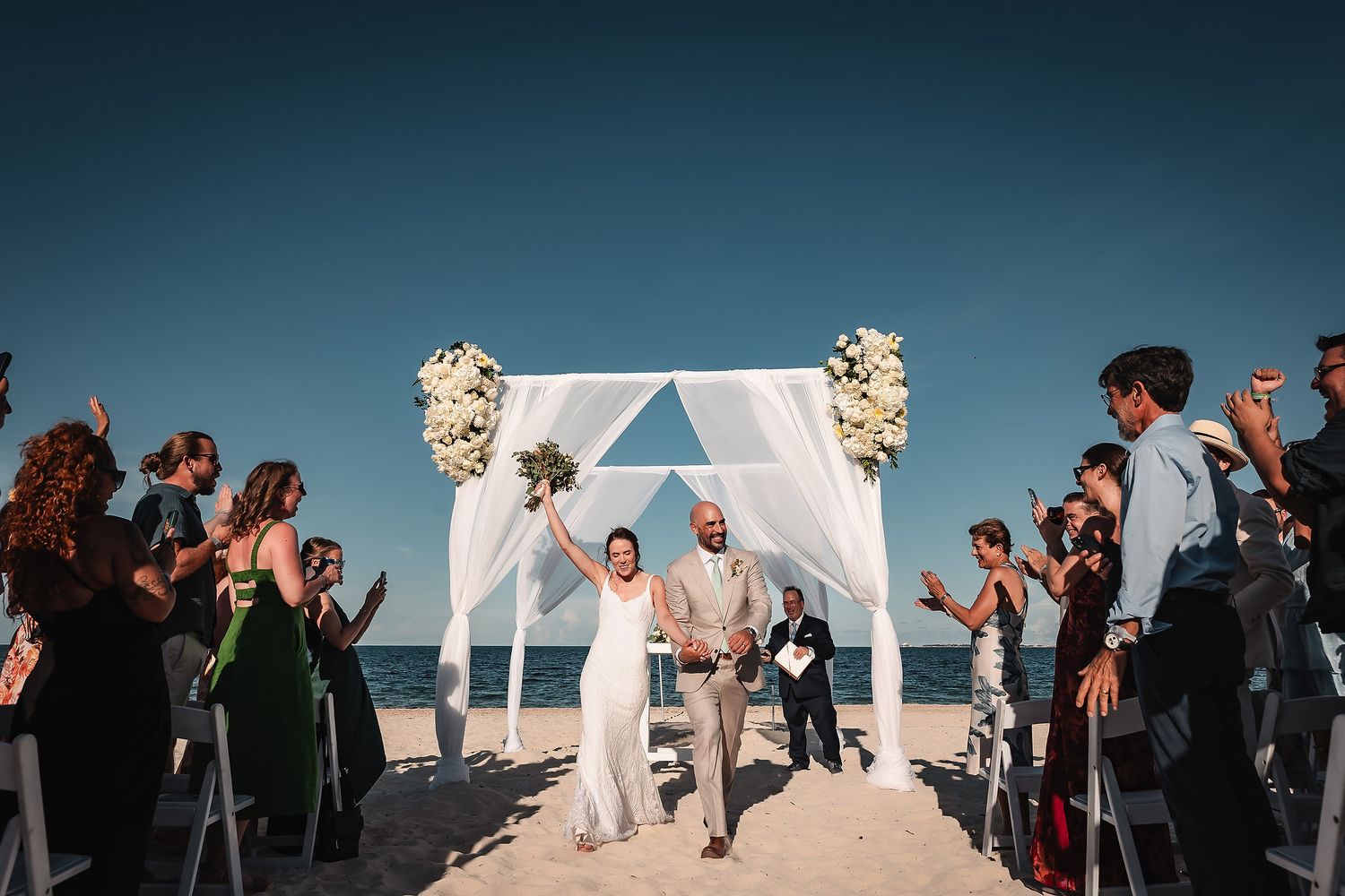 Wedding celebration on beach with guests cheering as ceremony concludes under white floral arch.