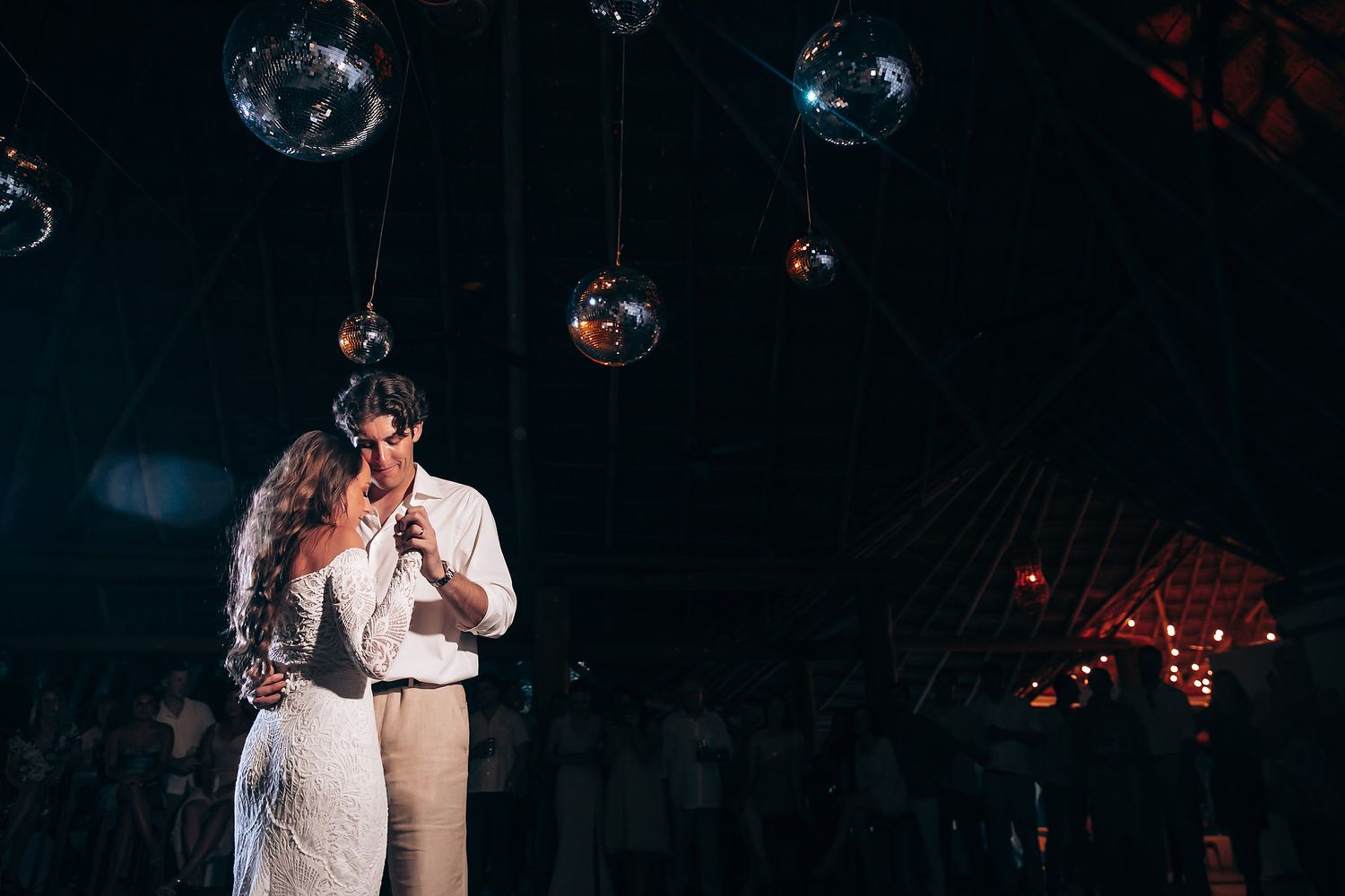 Newlyweds dance romantically beneath glowing disco balls in a dark reception venue.