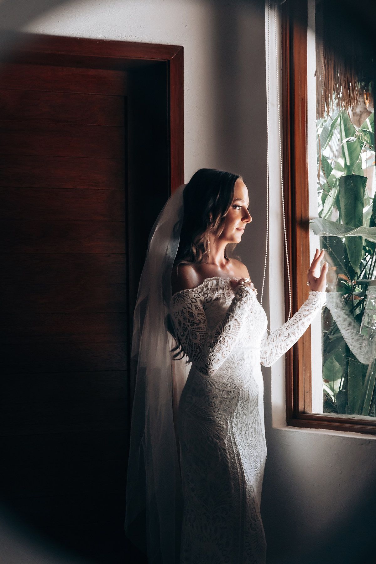 Bride in white lace dress poses by window with natural light streaming in.