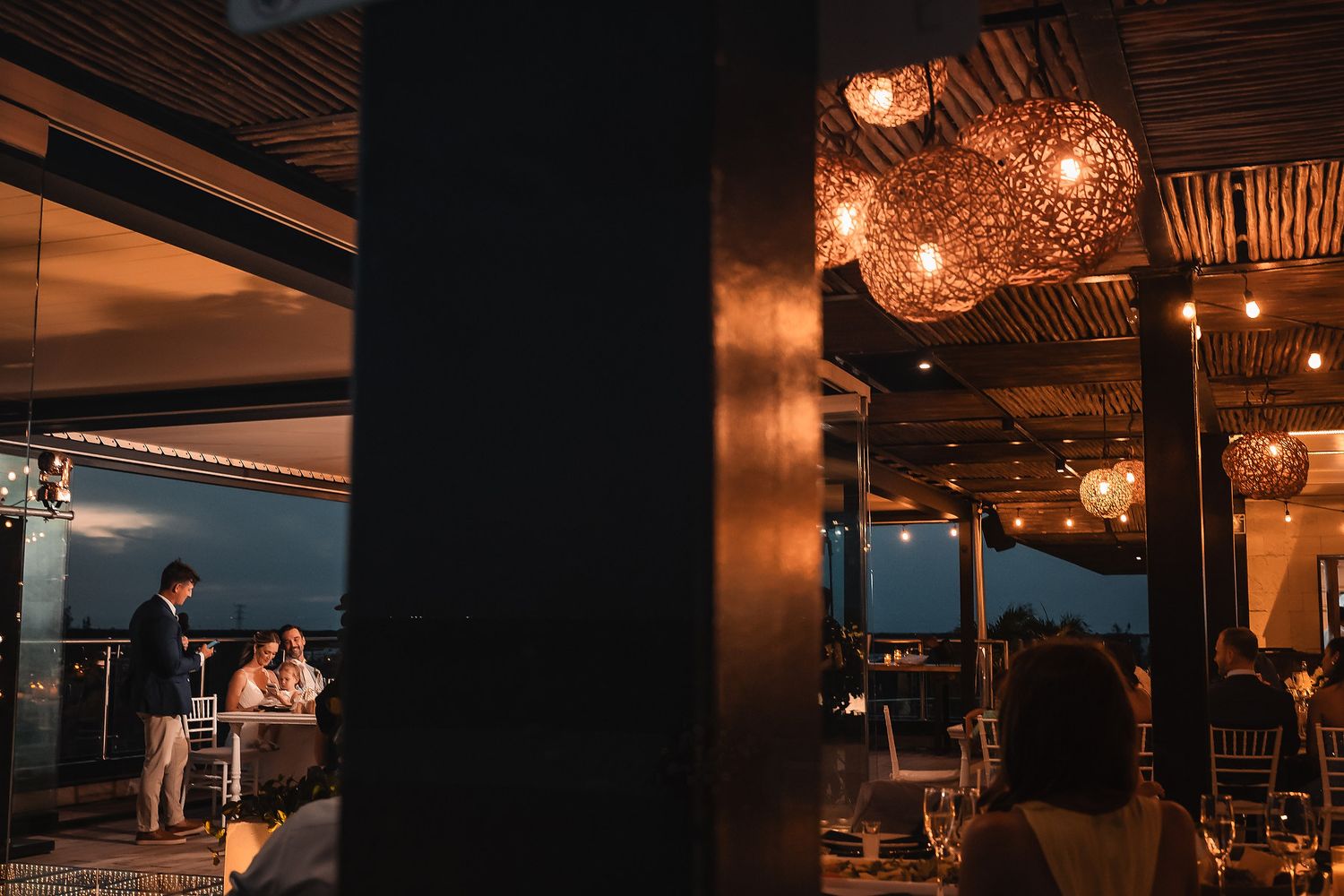 Romantic evening wedding reception venue with warm lighting and coastal views through glass windows.
