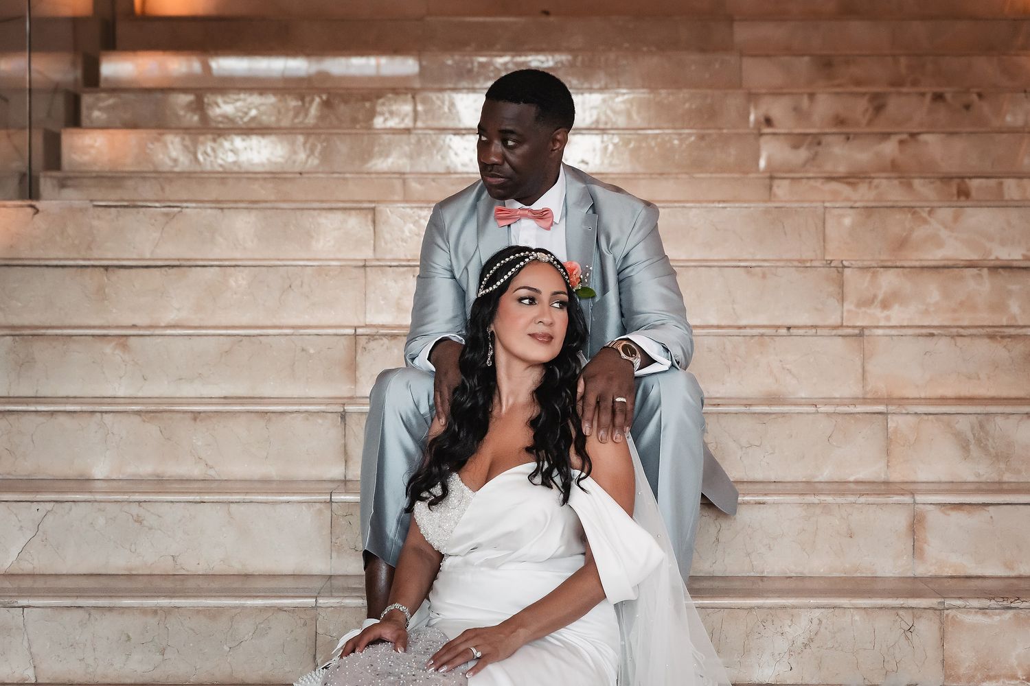 Newlyweds in formal attire pose together on elegant stone steps.