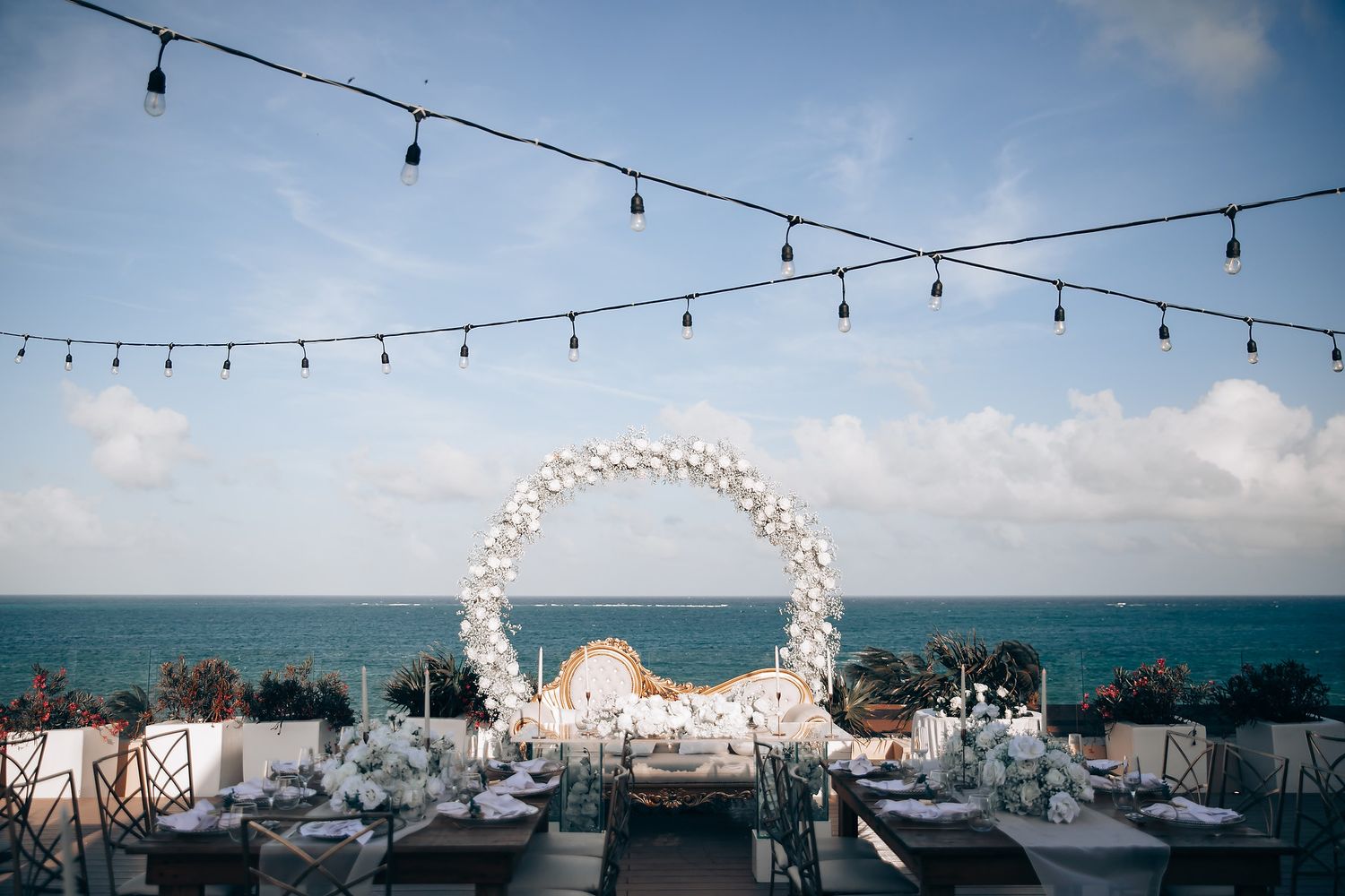 String lights hang across an outdoor venue with ocean views and blue skies in the background.