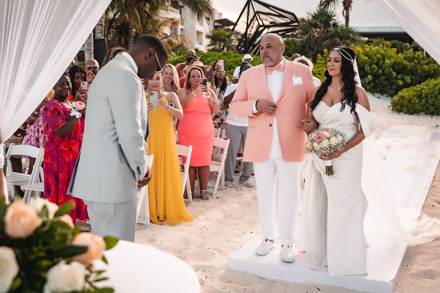 Beachside wedding ceremony setup with white drapes and colorful guest attire.
