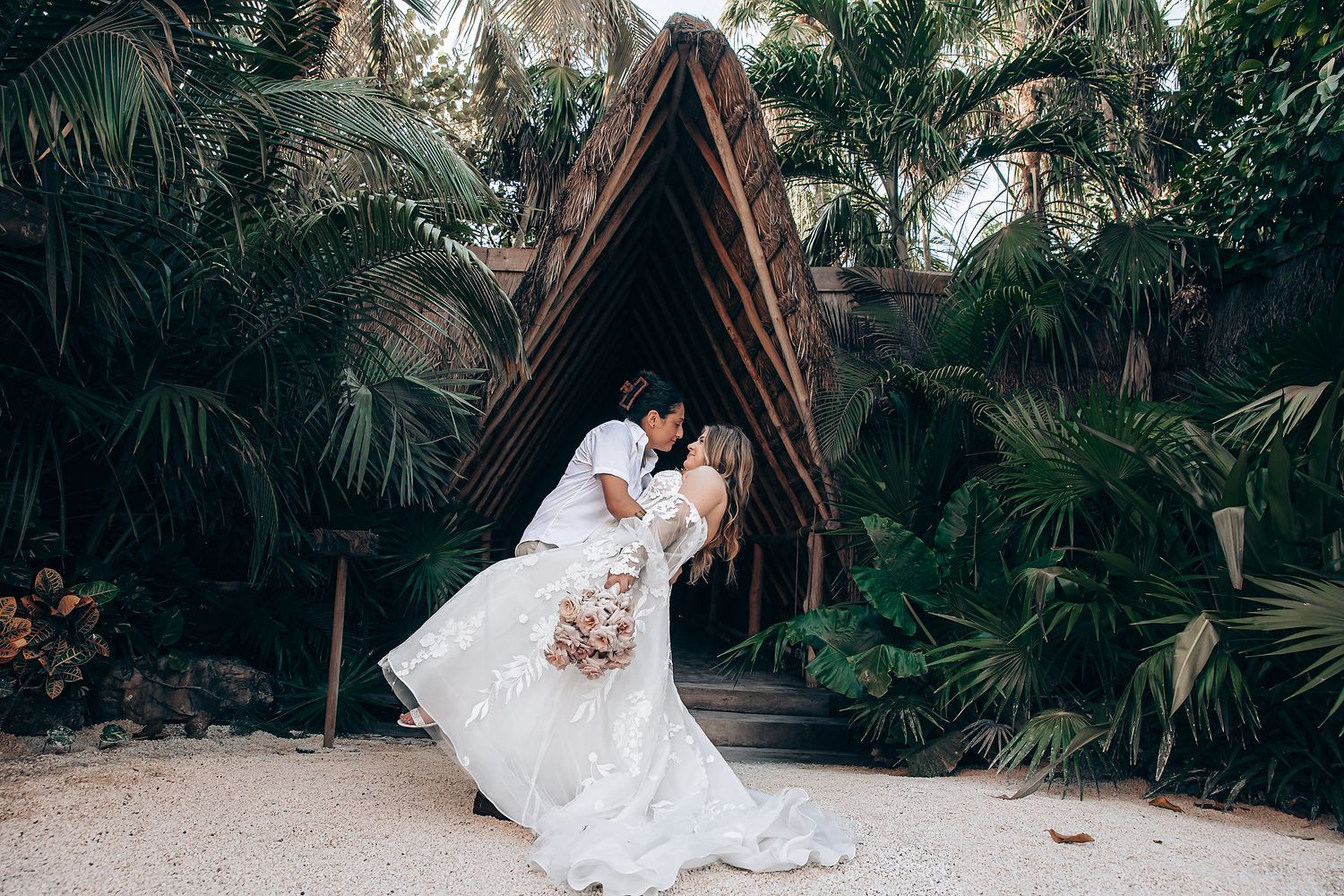 A couple shares a romantic dip pose under a wooden arch surrounded by tropical plants.