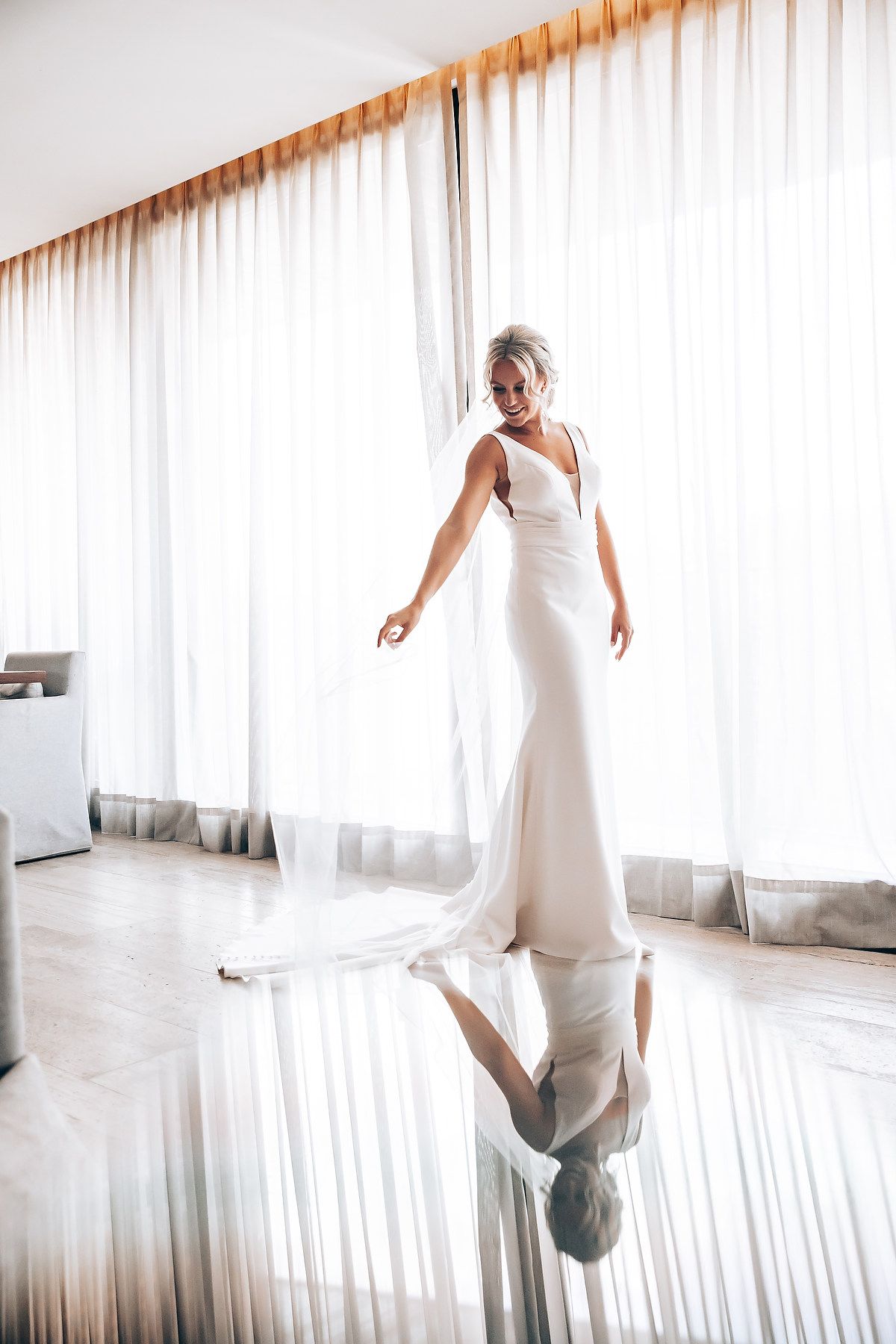Bridal portrait in sleek white dress reflected on glossy floor with window light.