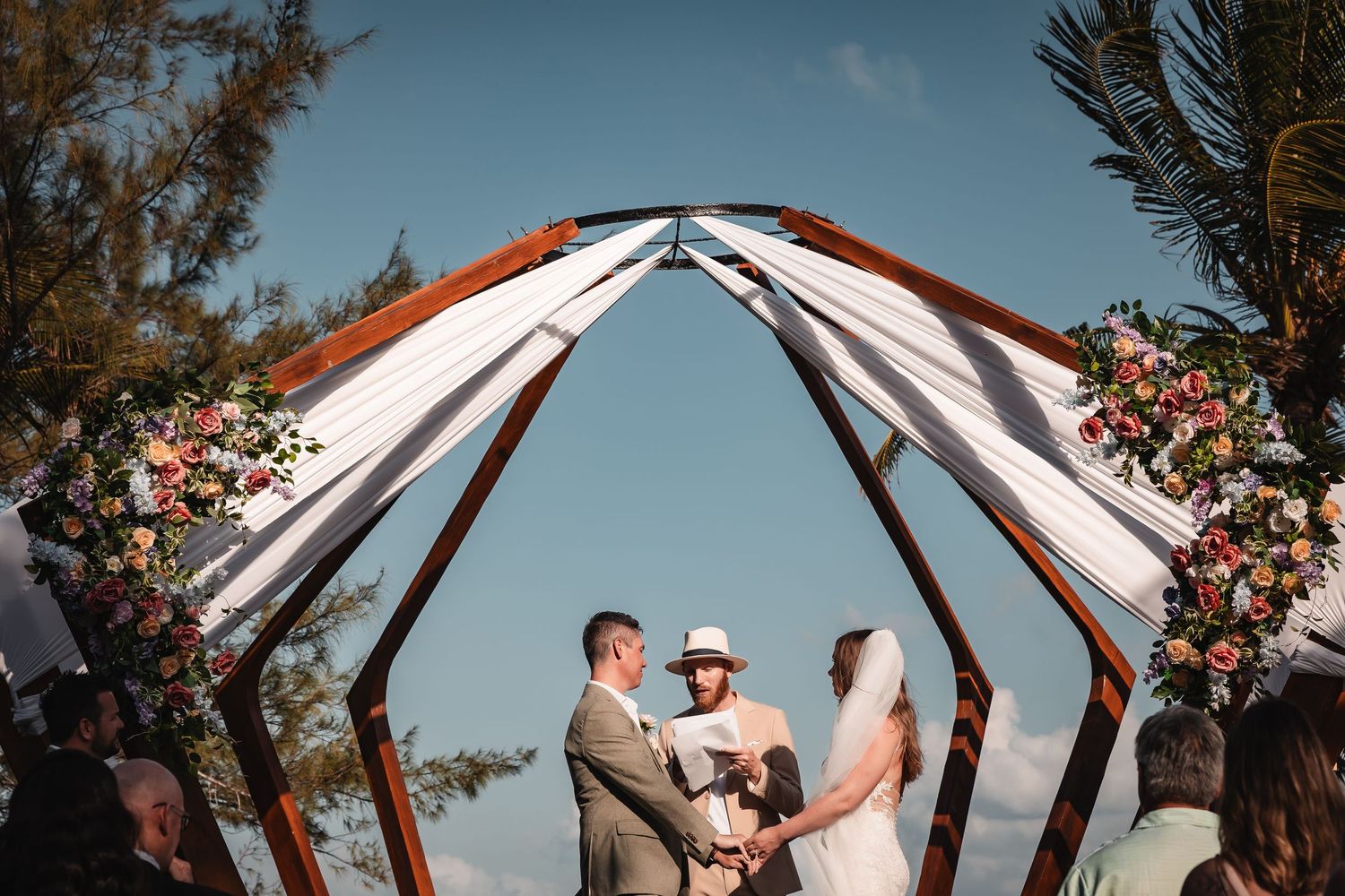 Wedding ceremony under geometric wooden arch with white fabric and tropical flowers on beach.