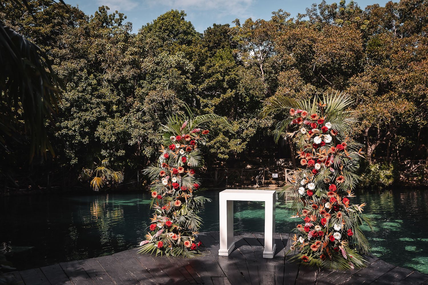 Wedding ceremony space decorated with red floral arrangements and white chairs in a tropical garden setting.